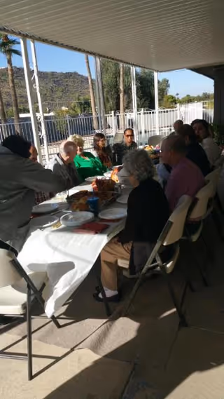 A group of elderly people sitting around a long table outdoors under a covered patio, enjoying a meal together with plates and food on the table. The background shows a fenced area with palm trees and hills in the distance.