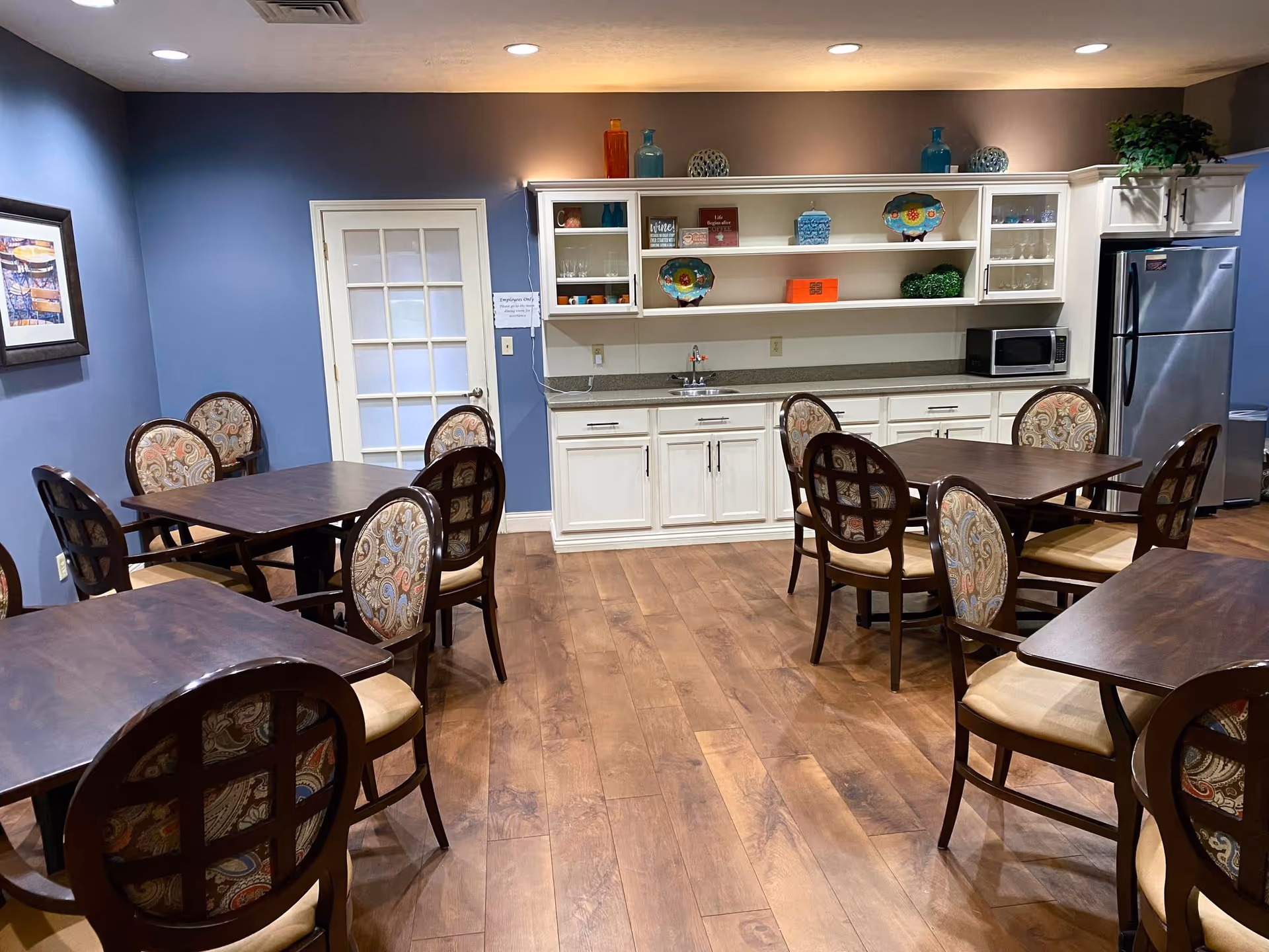 Communal dining room with several wooden tables and upholstered chairs facing a kitchenette with white cabinets, open shelving, and a refrigerator.