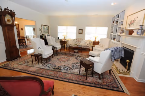 A spacious living room with a large patterned area rug covering the wooden floor. The room features several white upholstered armchairs arranged around a central coffee table, a floral-patterned sofa against the far wall, and a fireplace with a mantel decorated with framed artwork and vases. There is a grandfather clock near the entrance to an adjacent dining room, which is partially visible. The room is well-lit with natural light coming through two large windows.
