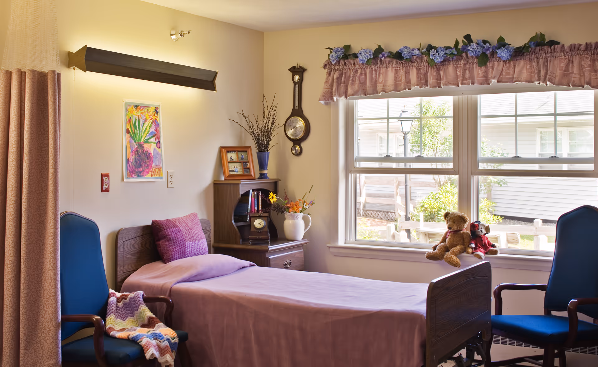 A cozy bedroom in a senior living facility with a single bed covered in a purple blanket and a matching pillow. There are two blue chairs on either side of the bed, one with a colorful knitted blanket draped over it. A wooden nightstand next to the bed holds a clock, a framed photo, a vase with flowers, and some books. Above the nightstand is a wall clock. The window has a floral valance and two stuffed animals, a teddy bear and a doll, sitting on the windowsill. Outside the window, there is a view of a garden and neighboring buildings.