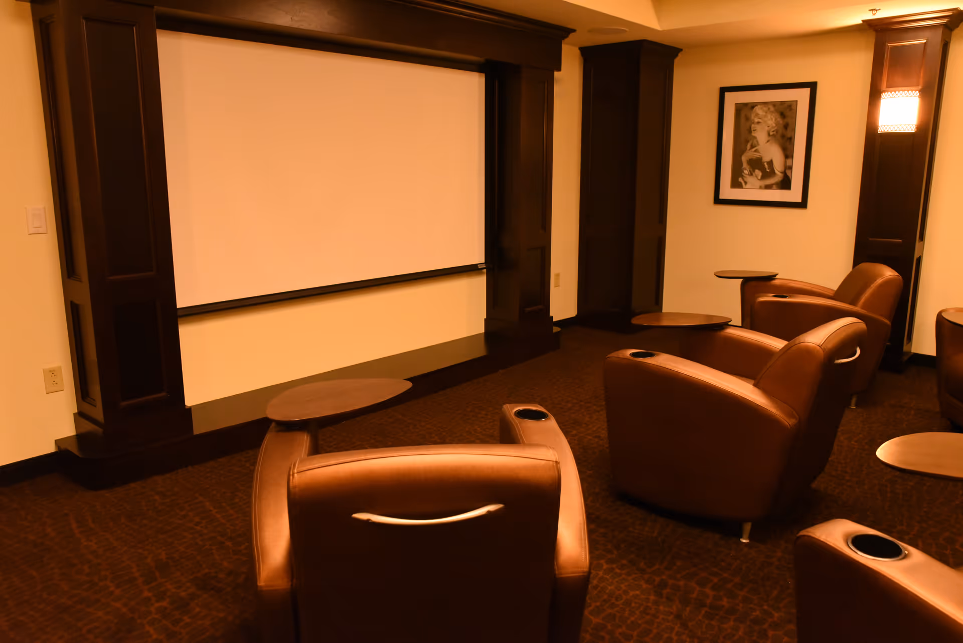 A cozy media room with several brown leather recliner chairs arranged facing a large blank projection screen. The room has dark wood paneling around the screen, a framed black and white photo on the wall, and warm lighting fixtures.
