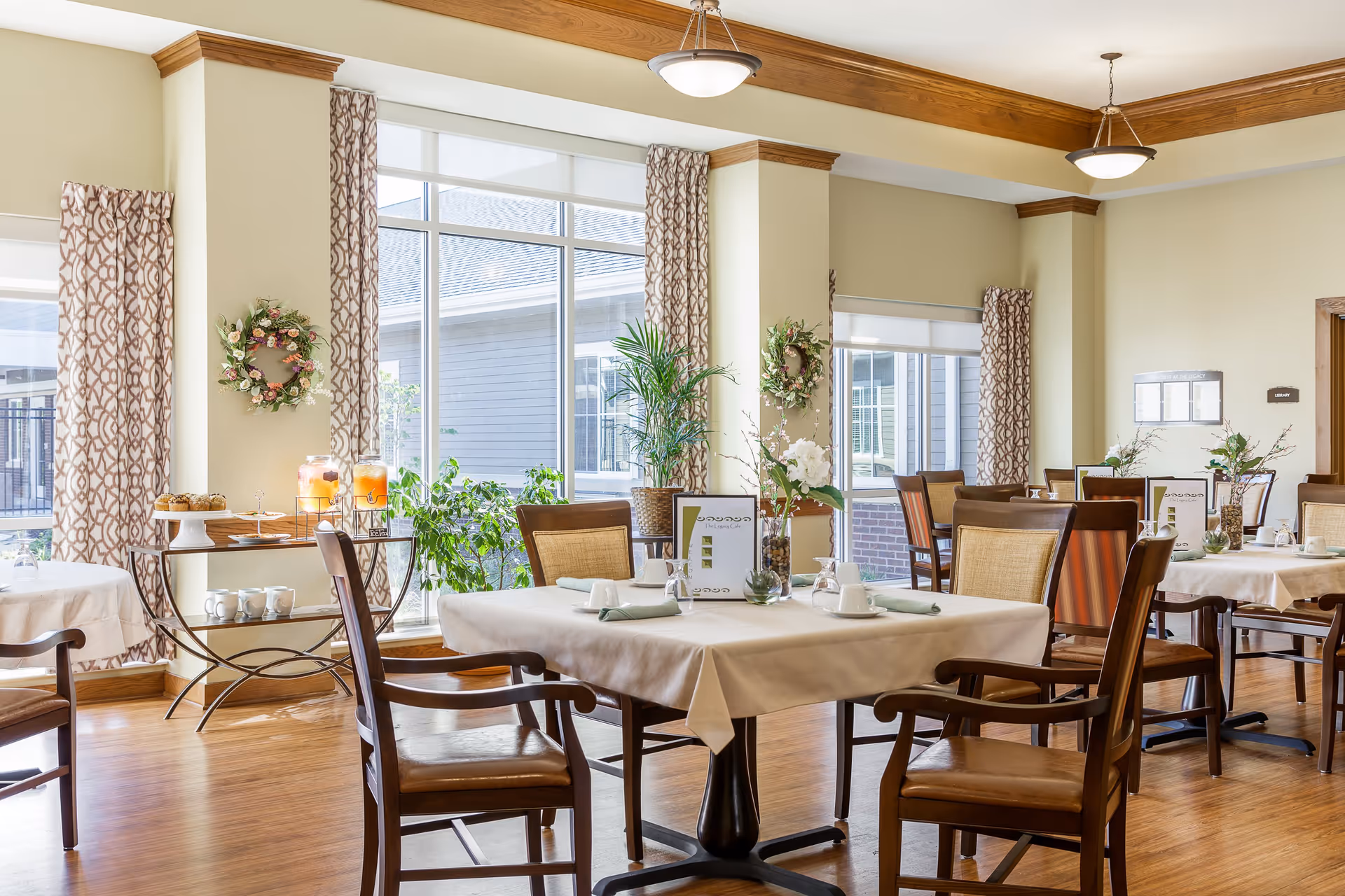 A bright and airy dining room with several tables covered in white tablecloths, set with cups, napkins, and small flower arrangements. Large windows with patterned curtains allow natural light to fill the room. There are wooden chairs around the tables and a small side table with beverages and pastries. The room features wooden crown molding and light fixtures hanging from the ceiling.