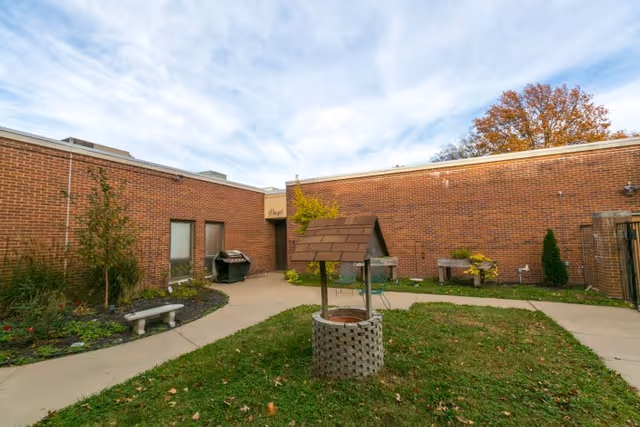 Outdoor courtyard area with a brick well structure in the center, surrounded by a concrete walkway. The courtyard is enclosed by brick walls of the building, with a door labeled 'Chapel' visible. There are some plants, a bench, and a barbecue grill along the walkway. The sky is partly cloudy.