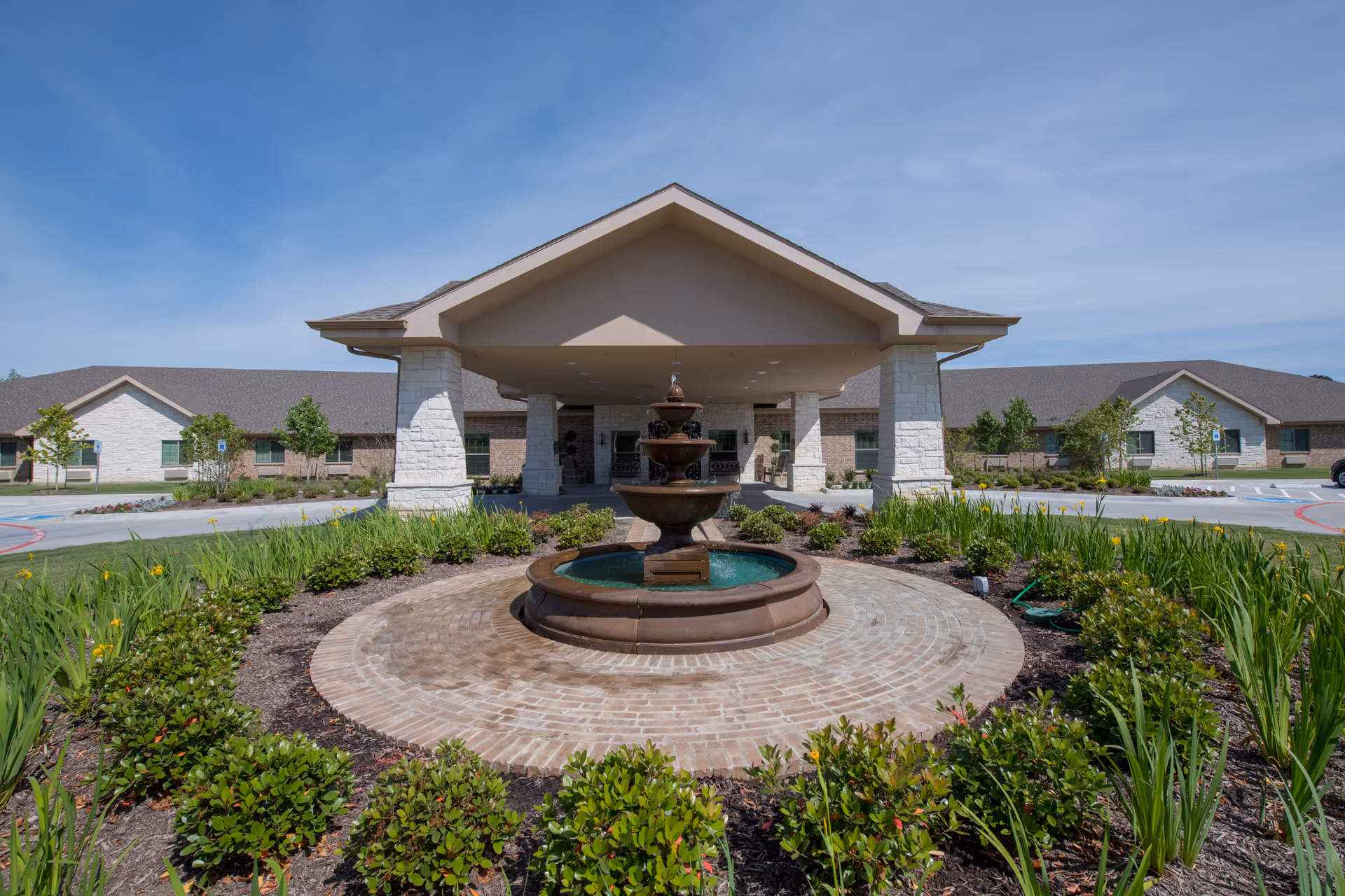 Front exterior view of RollingBrook Rehabilitation and Healthcare Center featuring a covered entrance with stone pillars, a central water fountain surrounded by landscaping, and a clear blue sky.