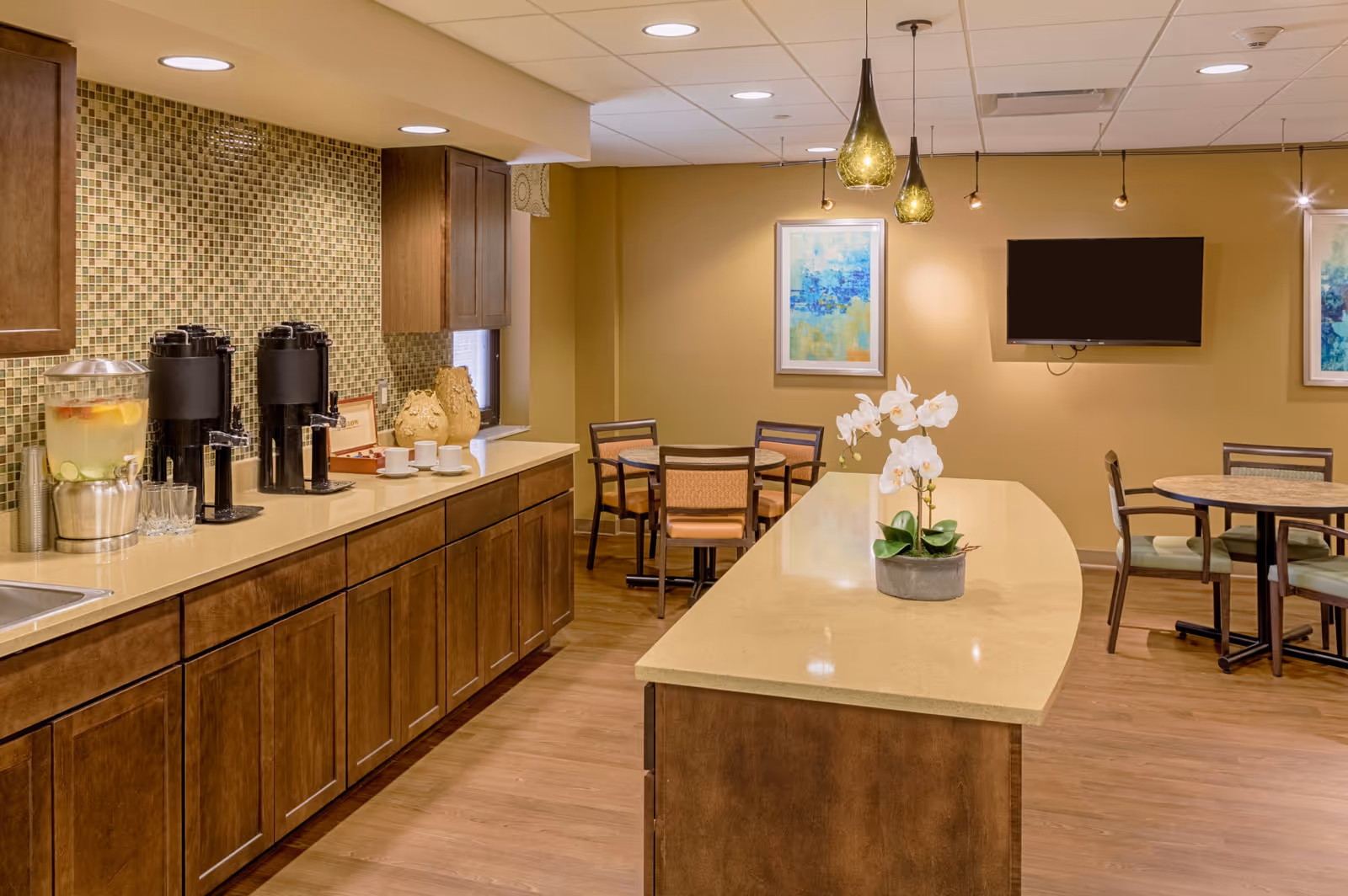 A cozy dining and refreshment area with wooden cabinets, a countertop with beverage dispensers, a water container with fruit slices, and a sink. There are round tables with chairs, a wall-mounted TV, framed abstract artwork, and pendant lights hanging from the ceiling. A white orchid plant is placed on the central island countertop.