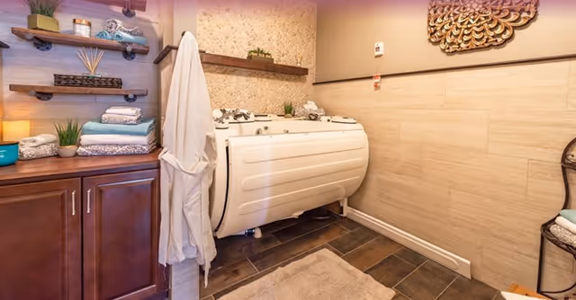 A well-lit bathroom featuring a white walk-in bathtub with safety handles, surrounded by beige tiled walls and dark wood flooring. To the left, there is a wooden cabinet with folded towels and decorative items on shelves above it. A white bathrobe hangs on a hook near the bathtub. On the right wall, a decorative golden wall art piece is displayed above a chair with folded towels.