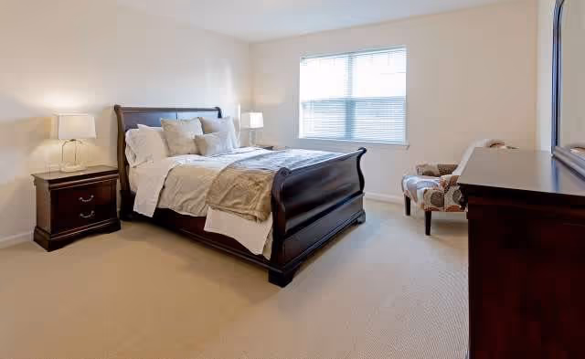 A well-lit bedroom with a large wooden bed featuring white and beige bedding, two matching wooden nightstands with lamps, a patterned armchair near a window with blinds, and a wooden dresser with a mirror above it.