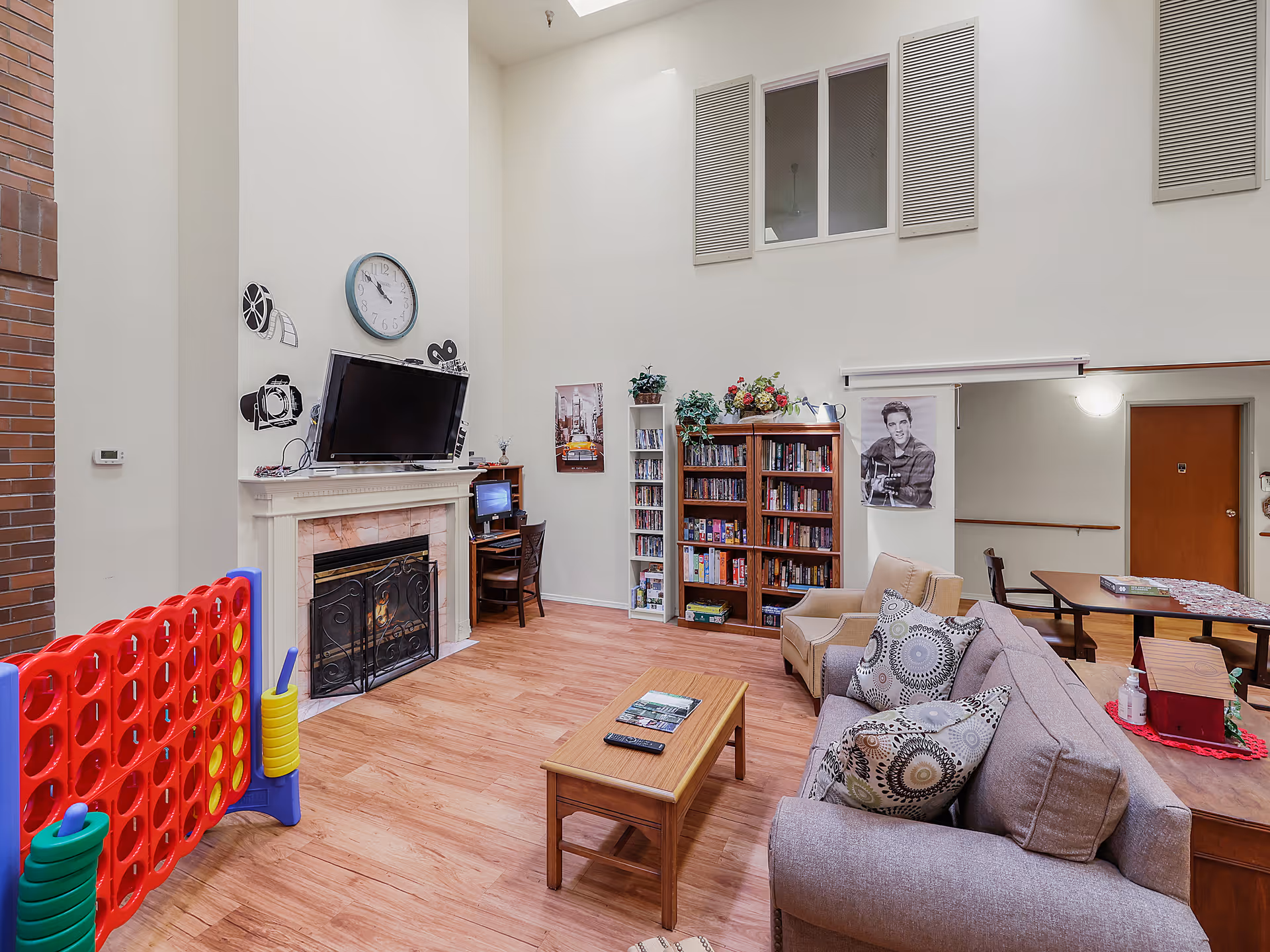 A cozy living room area with a fireplace, a wall-mounted TV above it, and a large Connect Four game on the left. There is a wooden coffee table in front of a gray couch with patterned pillows. A bookshelf filled with books and board games is against the far wall, along with a small desk and chair. The room has high ceilings with windows near the top and light wood flooring.