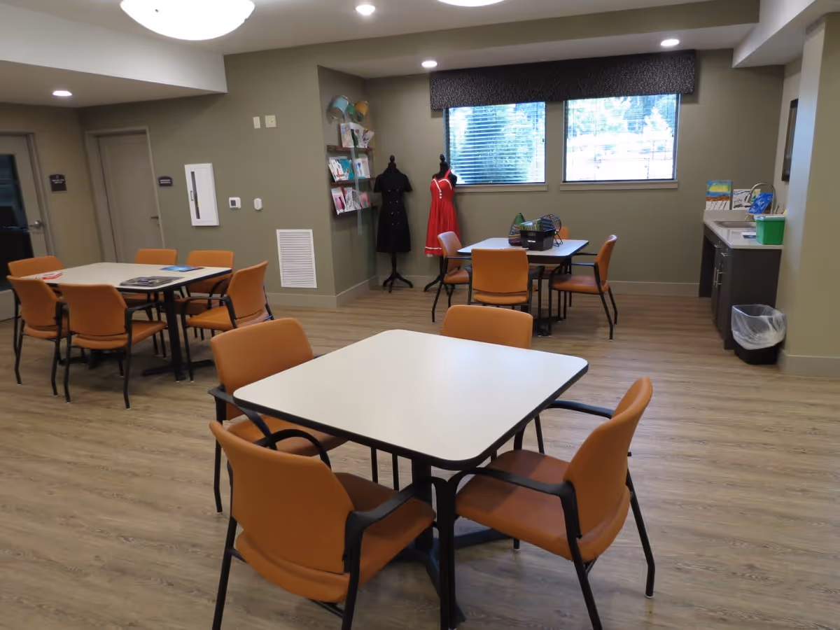 A well-lit room with three square tables, each surrounded by four orange chairs. The room has wood flooring, two windows with blinds, a small counter with a sink and a trash bin, and two dress mannequins displaying a black dress and a red dress. There are also shelves with books and magazines on the wall.