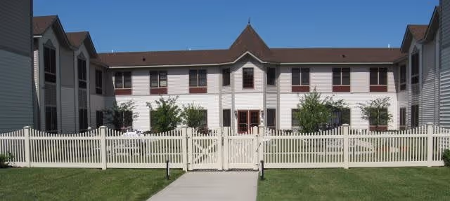 Two-story white building with a brown roof behind a white picket fence and central entrance under a clear blue sky.