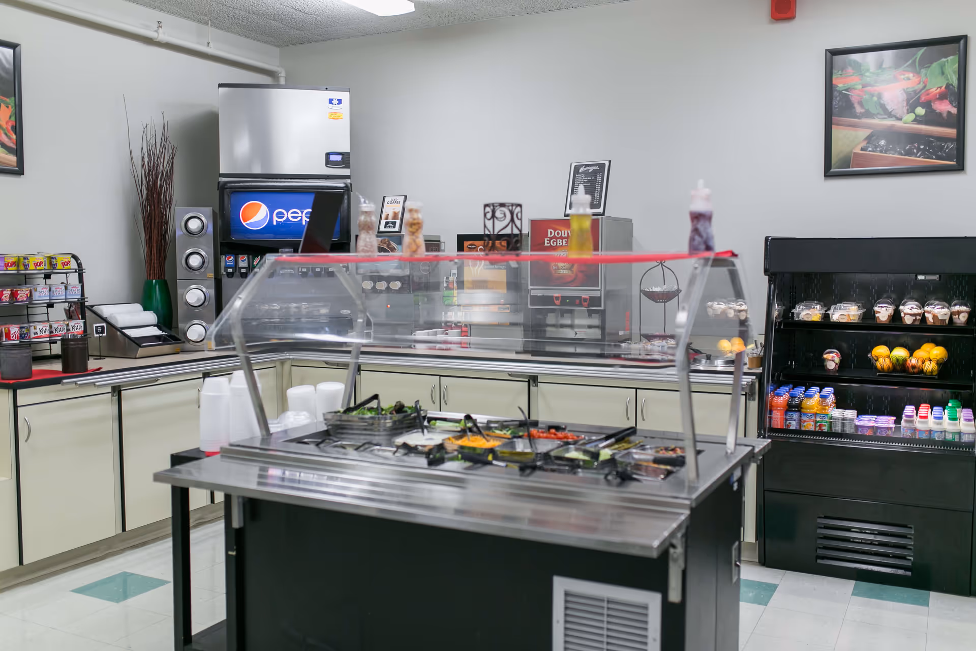 Interior view of a cafeteria or dining area with a salad bar in the center, a Pepsi soda machine, coffee dispensers, and a refrigerated display case with fruit, yogurt, and beverages. The room has light-colored walls and cabinets, with framed pictures on the walls.