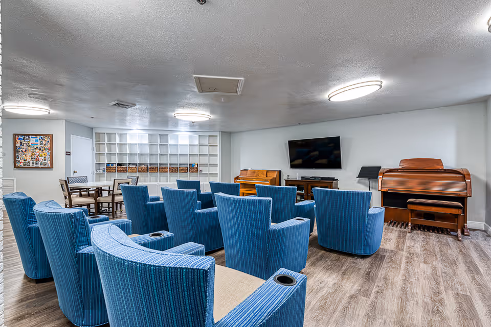 A common area in a senior living facility with multiple blue upholstered chairs arranged in rows facing a wall-mounted flat screen TV. There is a wooden piano and an organ to the right of the TV. In the background, there are tables and chairs near a wall with built-in shelves containing baskets. The room has wood flooring and white walls with ceiling lights.