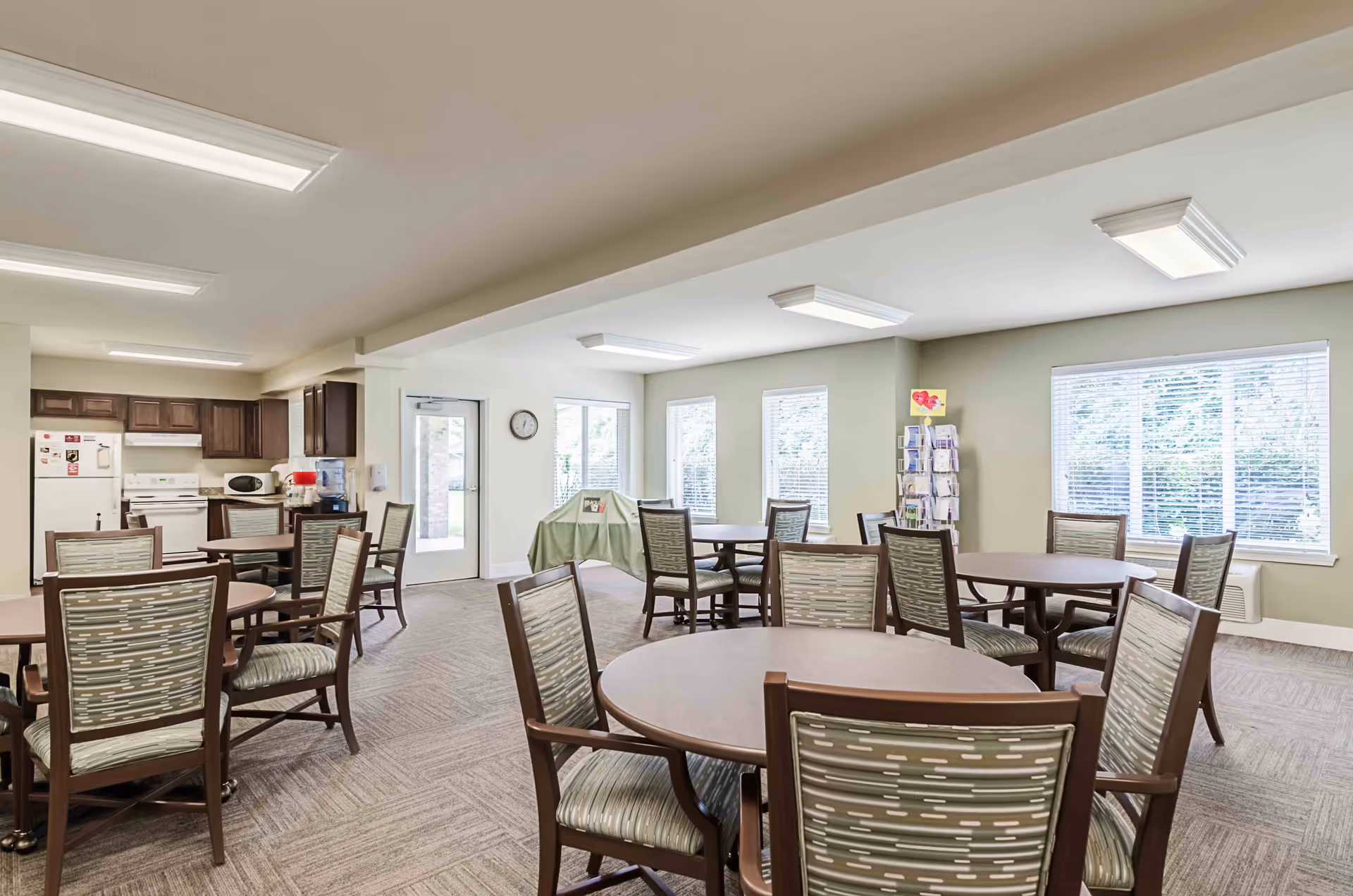 A bright dining room with several round tables and chairs arranged neatly. The room has large windows with blinds allowing natural light to fill the space. In the background, there is a kitchen area with wooden cabinets, a white refrigerator, stove, microwave, and a water dispenser. The walls are painted light green, and the floor is carpeted.