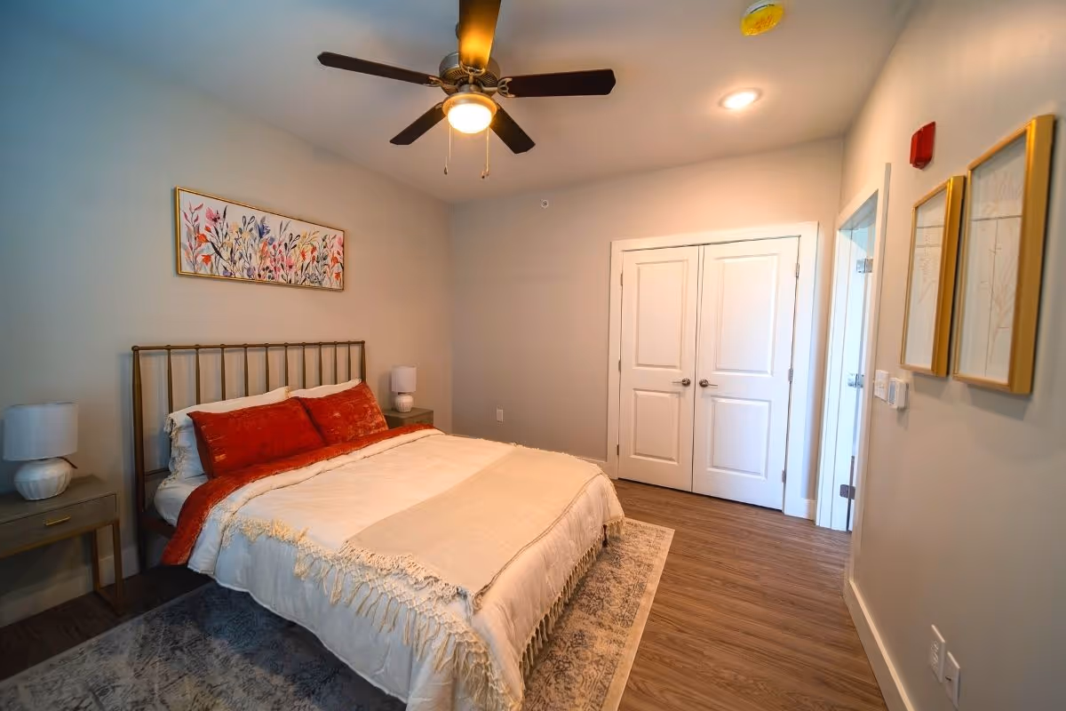 A cozy bedroom with a double bed featuring a metal headboard, white and red bedding, and two bedside tables each with a lamp. The room has wooden flooring, a ceiling fan with light, two framed artworks on the wall, and a closed double-door closet.