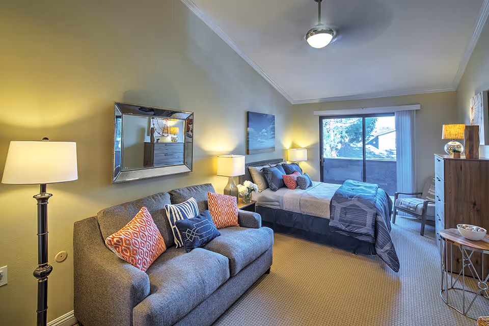 A cozy senior living bedroom featuring a bed with blue and beige bedding, a gray sofa with colorful pillows, a floor lamp, two bedside tables with lamps, a wooden dresser, a chair, and a sliding glass door leading to an outdoor balcony.