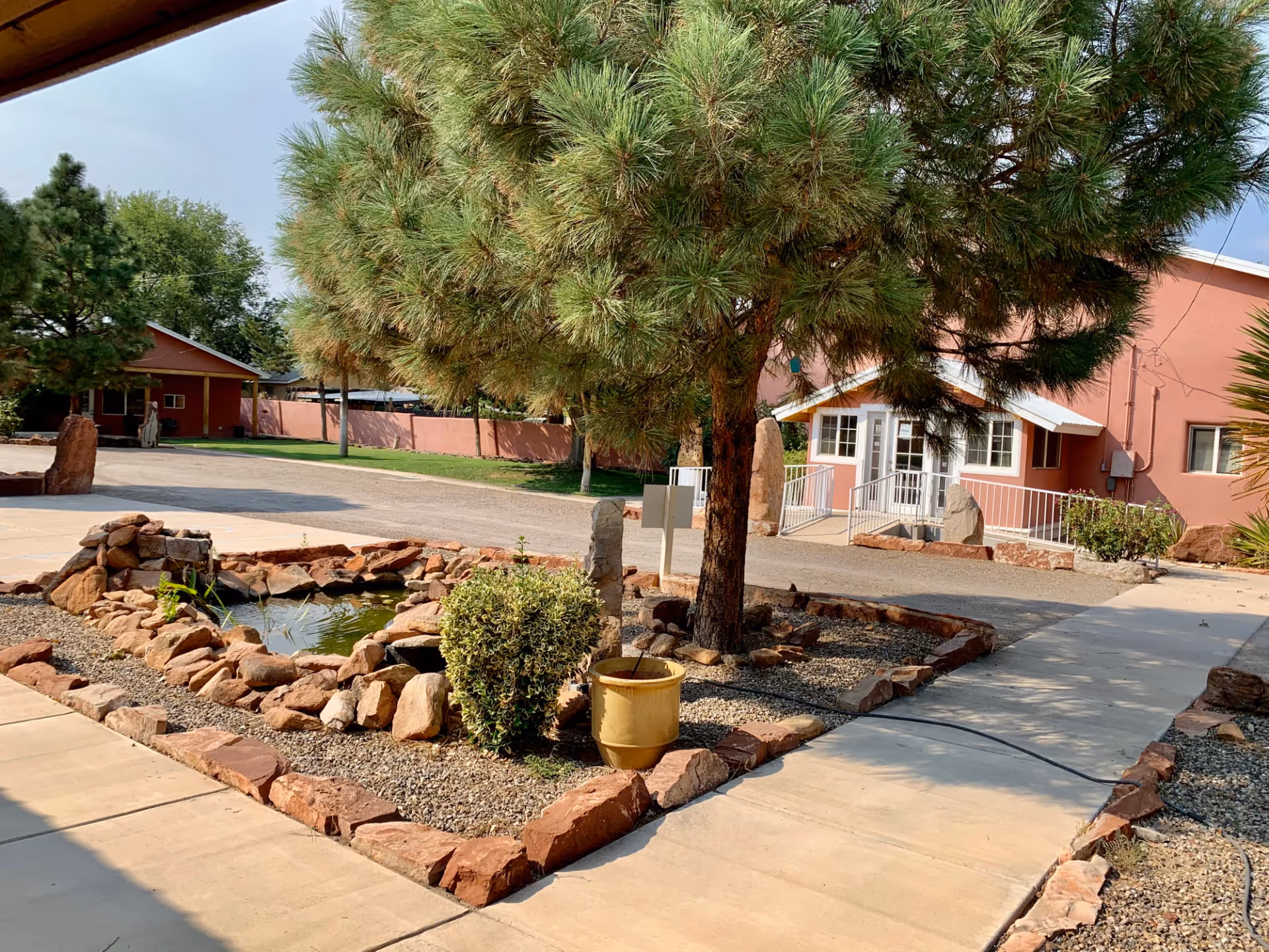 Outdoor view of Casa de Rosa Assisted Living showing a landscaped area with a small pond surrounded by rocks, a tree, and some bushes. There is a paved walkway leading to a pink building with white trim and a ramp entrance. The sky is clear and the area is well-maintained.