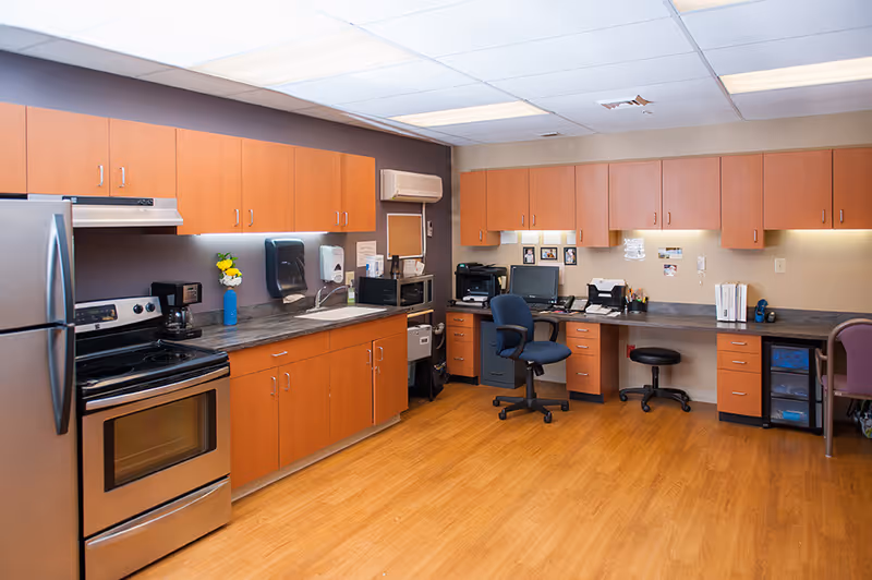 A well-lit room featuring a kitchen area with wooden cabinets, a stainless steel refrigerator, stove, and coffee maker on the left side. The right side has a long desk with office equipment including a computer, printer, and chairs, indicating a combined kitchen and office workspace.