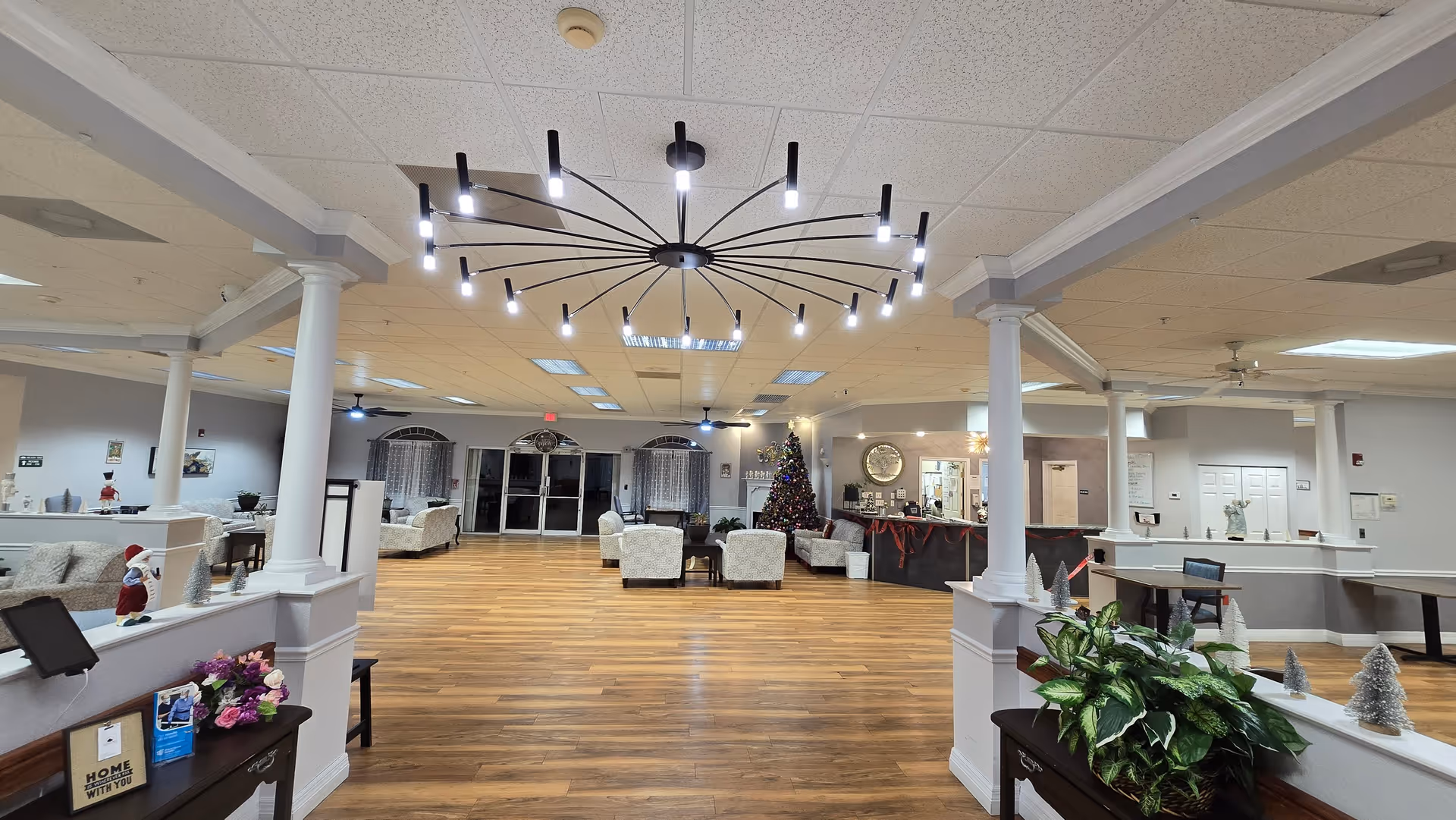 Spacious assisted living facility common area with wooden flooring, white pillars, and modern ceiling lights. The room is furnished with multiple seating arrangements including sofas and armchairs. A decorated Christmas tree is visible near the reception desk, which is adorned with holiday garlands. There are plants and small decorative items on tables and ledges throughout the space.