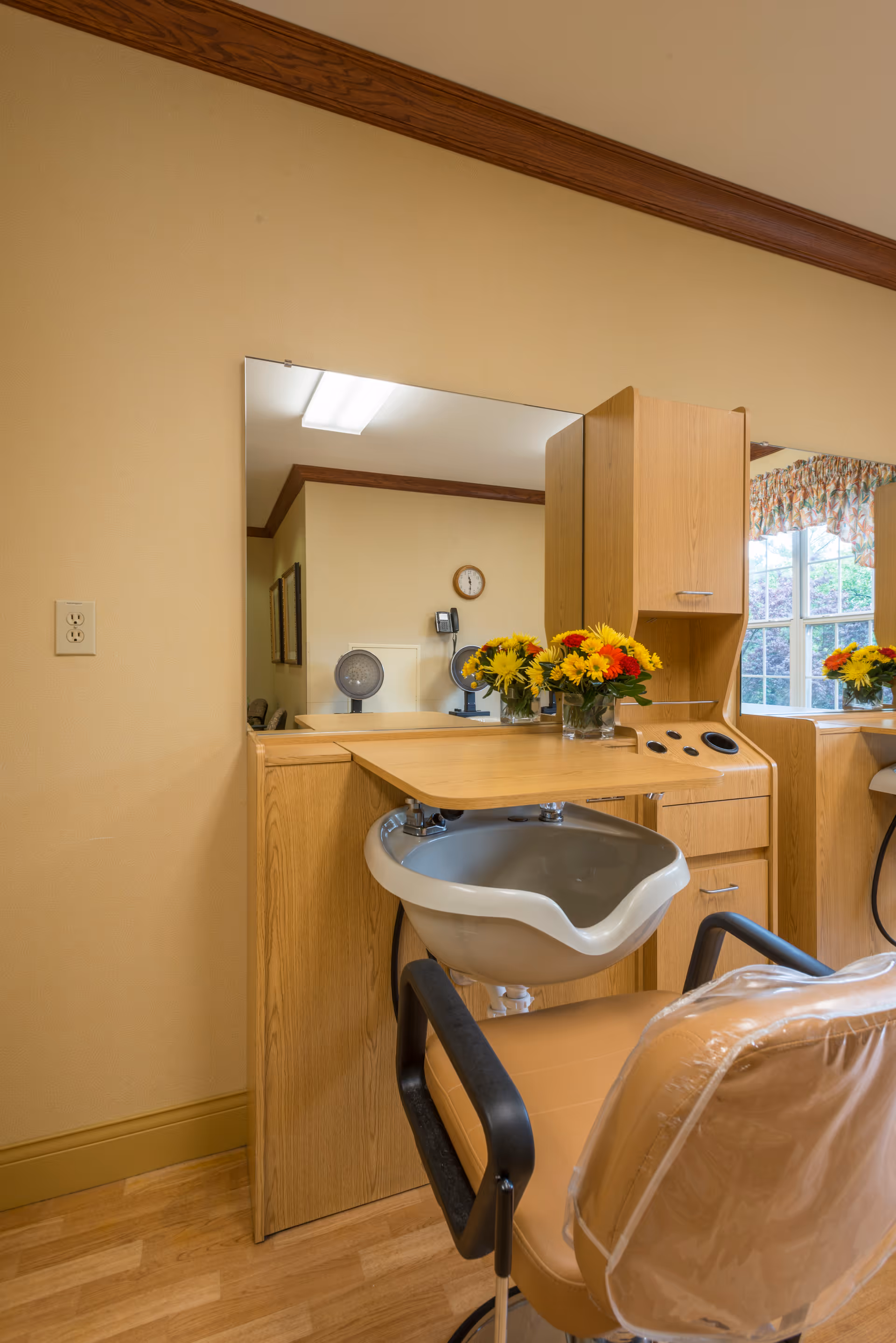 Interior view of a hair salon station in a senior living facility, featuring a beige salon chair with black armrests, a white shampoo basin, a wooden counter with drawers, a large mirror, and a vase of yellow and red flowers. The room has beige walls with wooden trim and a window with floral curtains.
