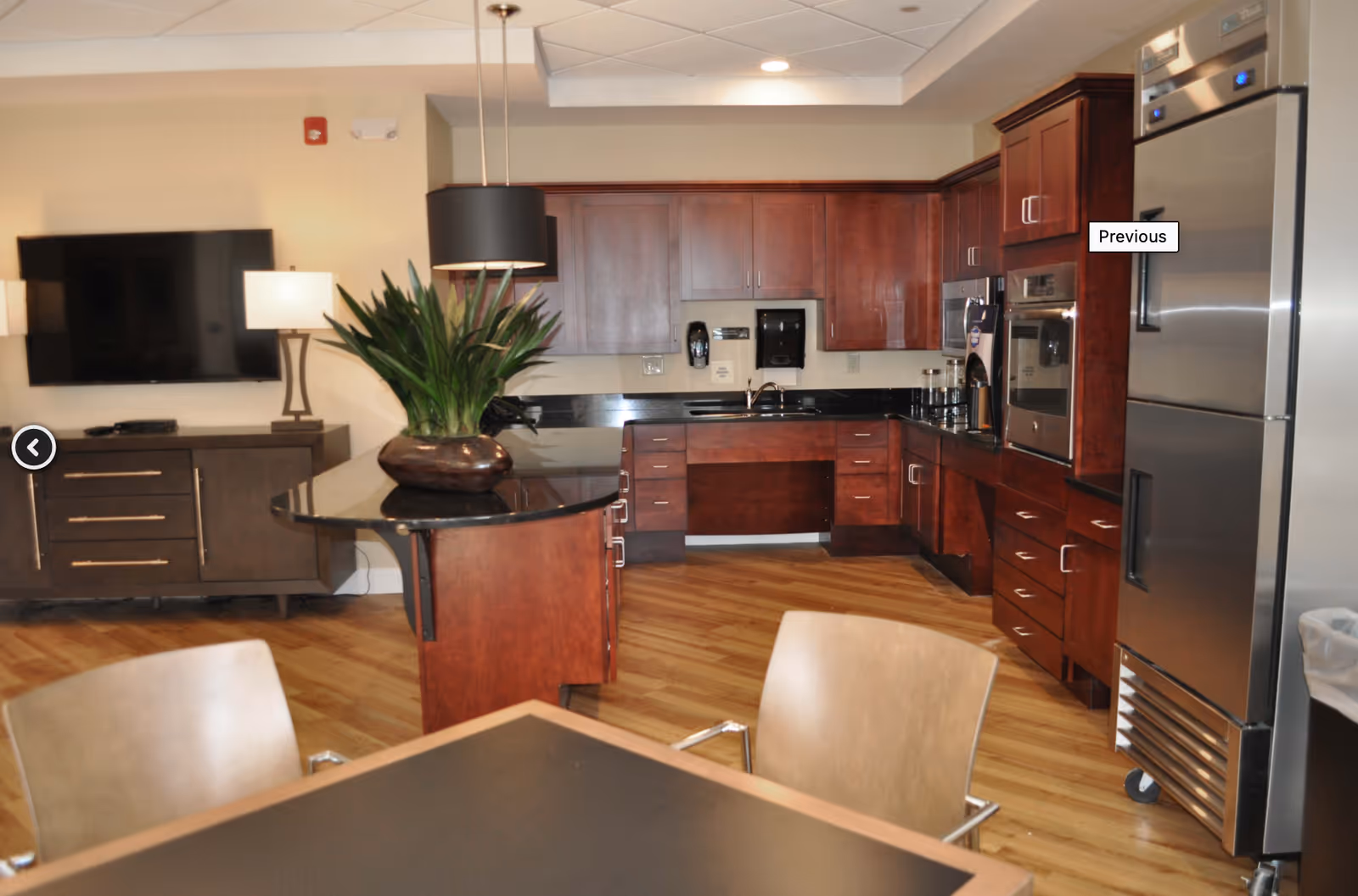 Interior view of a modern kitchen area in an assisted living facility featuring wooden cabinets, a black countertop island with a plant centerpiece, stainless steel refrigerator, built-in oven, microwave, and a dining table with chairs in the foreground. A flat-screen TV and a lamp are visible on a cabinet against the wall.