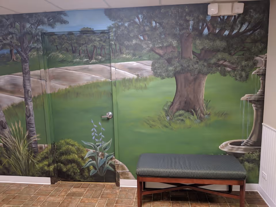 Indoor area with a mural painted on the wall depicting a green landscape with trees, bushes, a stone pathway, and a water fountain. There is a door camouflaged within the mural and a cushioned bench with wooden legs placed against the wall.