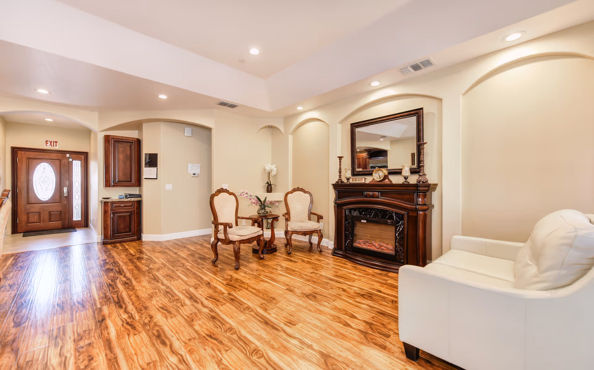 A cozy senior living room with polished wooden floors, two vintage-style armchairs with beige upholstery, a small round table with a flower arrangement, a white cushioned sofa, and an elegant dark wooden electric fireplace with a mirror above it. The room has soft beige walls with recessed lighting and an entrance door with an exit sign above it.