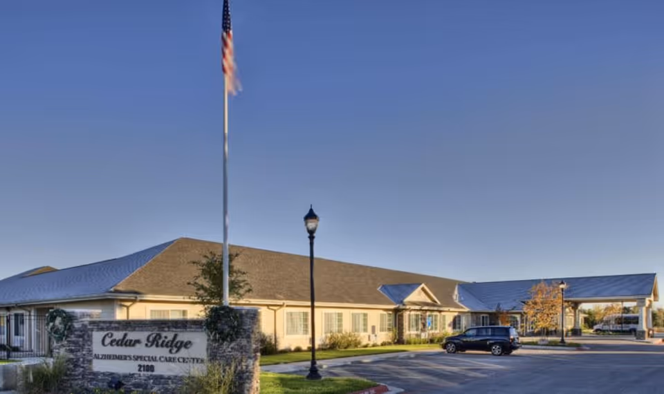 Exterior view of Cedar Ridge Alzheimer's Special Care Center building with a flagpole and street lamp in front, a parking lot with a black SUV, and clear blue sky.