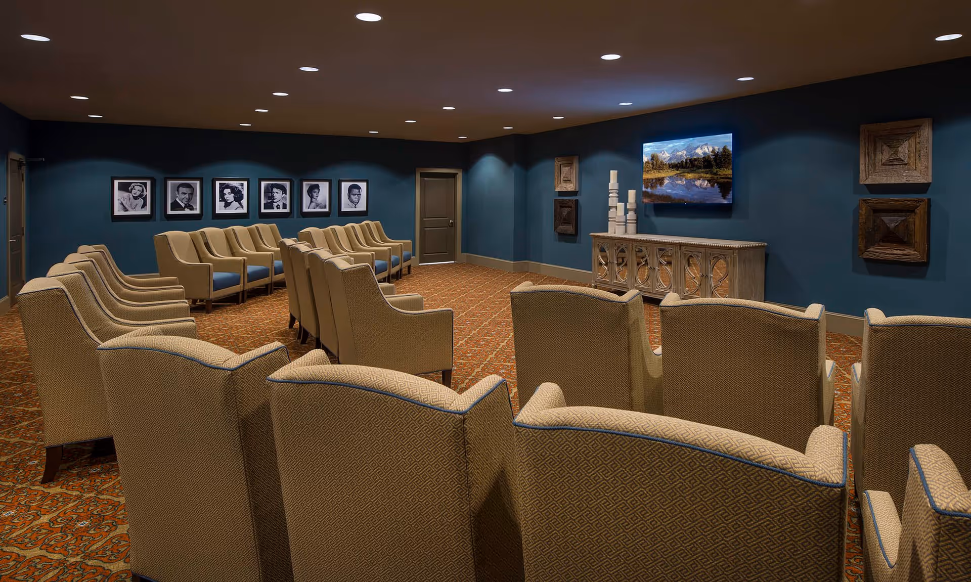 A cozy room with multiple beige upholstered chairs arranged in rows facing a wall-mounted television displaying a nature scene. The walls are painted dark blue and decorated with framed black and white portraits and wooden art pieces. The carpet has a patterned design, and the ceiling has recessed lighting.