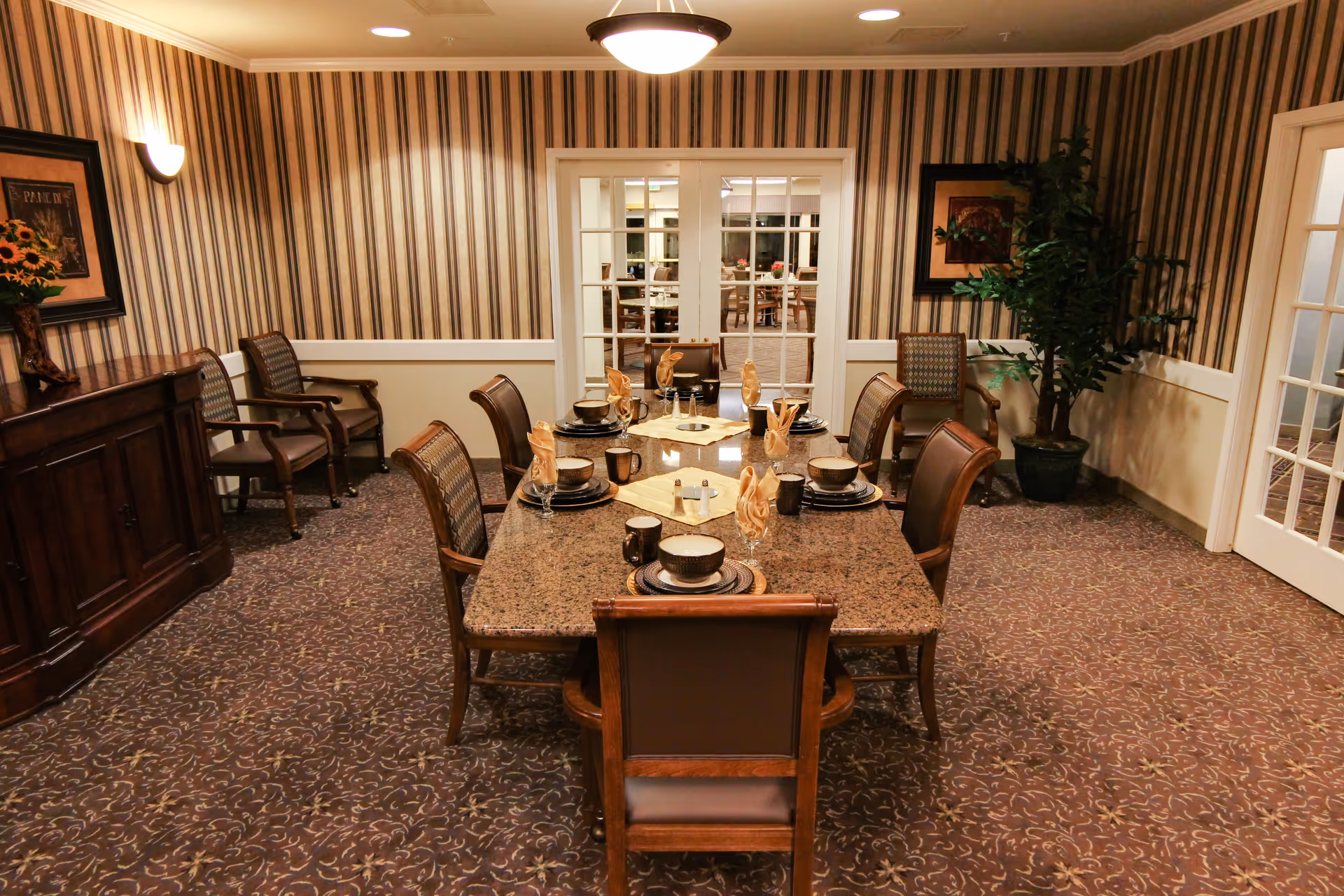 A formal dining room with a rectangular granite table set for six people with plates, bowls, mugs, and folded napkins. The room has patterned carpet, striped wallpaper, and wooden chairs with cushioned seats. There are two sets of French doors, a wooden sideboard with a vase of sunflowers, framed artwork on the walls, and a large potted plant in the corner.
