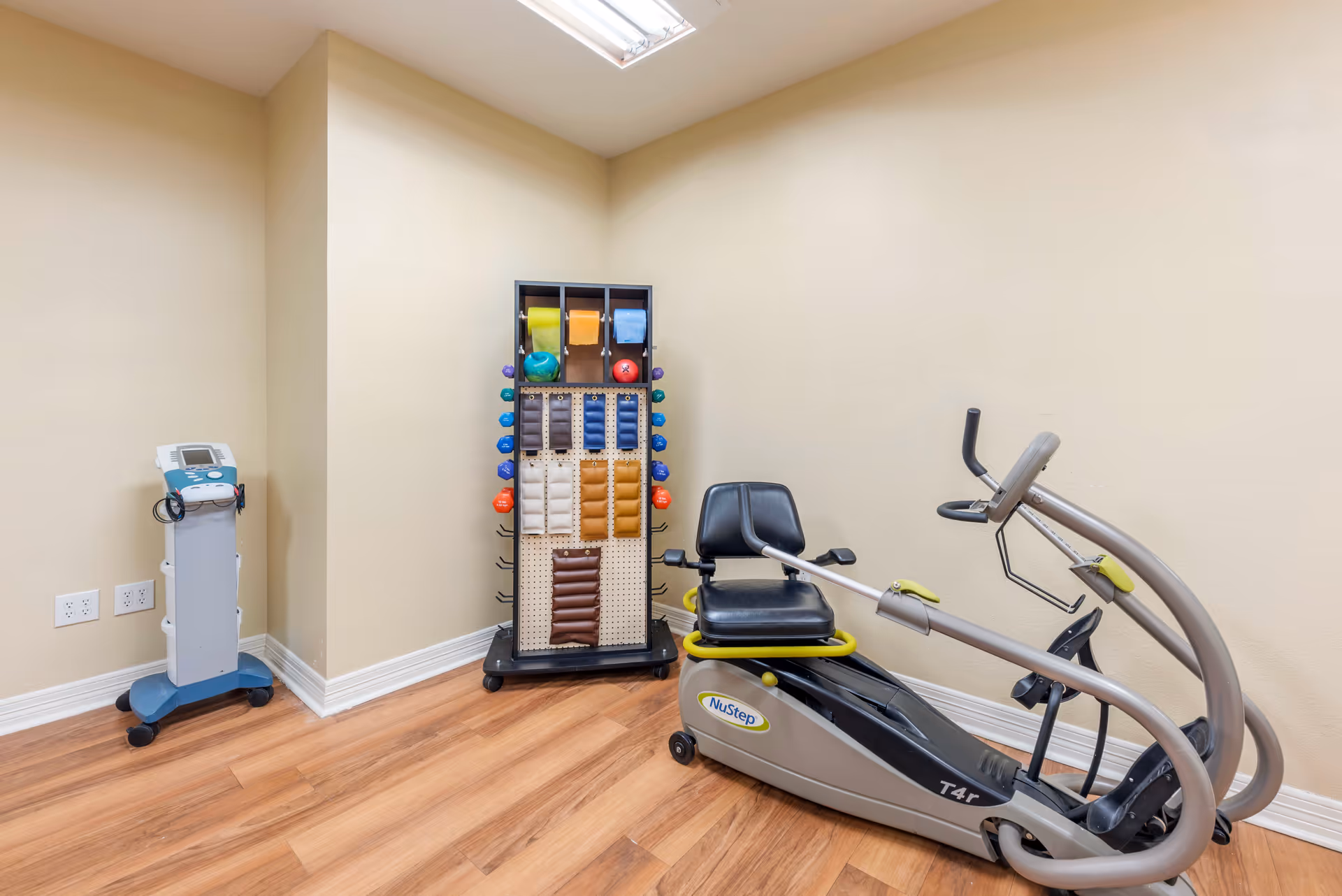A small exercise room with a NuStep T4r recumbent cross trainer, a rack holding various colored exercise bands and small dumbbells, and a medical device on a stand in the corner. The room has beige walls and wood flooring with a fluorescent light on the ceiling.