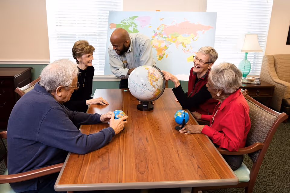 A group of four elderly people and one staff member sitting around a wooden table in a well-lit room. They are engaged in a geography activity with a large globe in the center of the table and two smaller globes held by two of the elderly individuals. A world map is displayed on the wall behind them, and the atmosphere appears friendly and interactive.