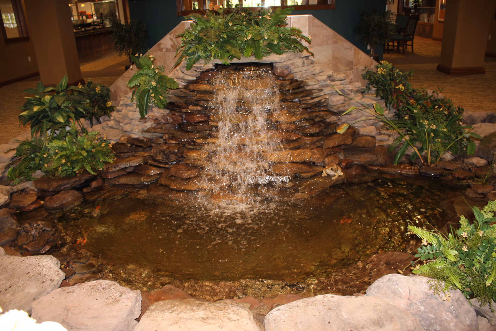 Indoor water feature with a small waterfall flowing over stacked stones into a pond surrounded by various green plants and rocks, located inside a building with carpeted floors and visible seating areas in the background.