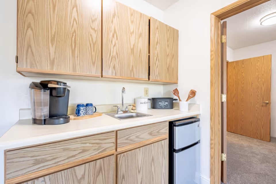 A small kitchenette area with light wood cabinets above and below a white countertop. On the countertop, there is a black coffee maker, two blue patterned mugs on a wooden tray, a small sink with a faucet, a white container labeled 'cookie', a black container labeled 'bread bin', and a white utensil holder with wooden spoons. To the right, there is a small silver refrigerator. An open doorway leads to a carpeted room with wooden doors.