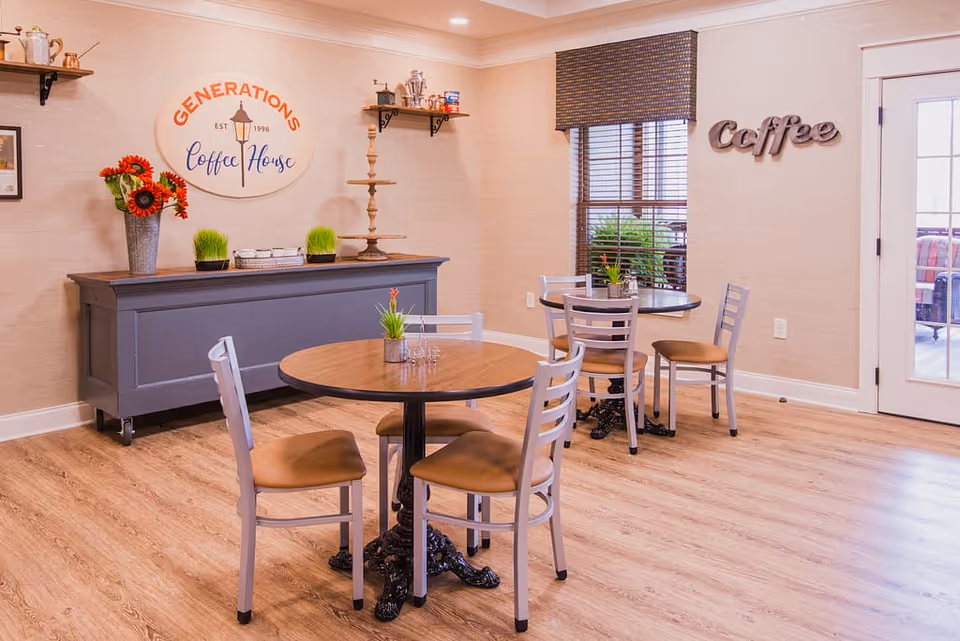 A cozy coffee house area with two round wooden tables, each surrounded by four chairs with tan cushions. A gray sideboard against the wall holds decorative items including a vase with red flowers and small green plants. Above the sideboard is a sign that reads 'Generations Coffee House EST 1996'. On the adjacent wall, the word 'Coffee' is displayed in large decorative letters. A window with blinds and a door with glass panes are visible, letting in natural light.