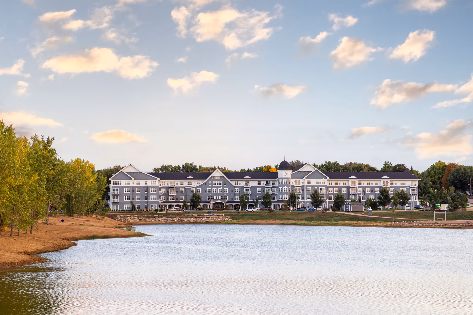 A large multi-story senior living building stretches along a lakeshore under a partly cloudy sky.