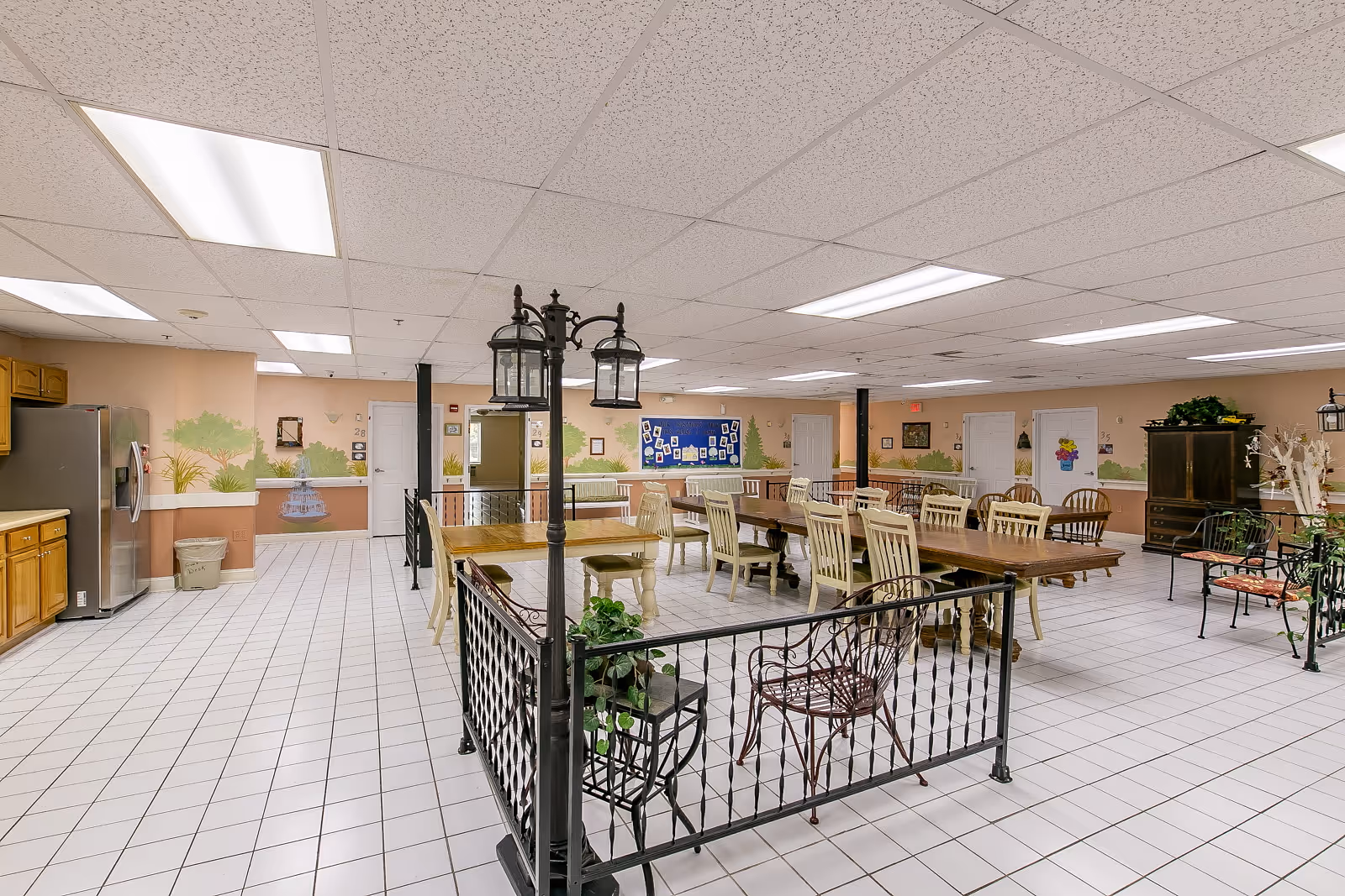 A spacious senior living facility dining room with multiple wooden tables and chairs arranged neatly. The room has a tiled floor, a drop ceiling with fluorescent lights, and a kitchen area with wooden cabinets and a stainless steel refrigerator on the left. Decorative elements include a black metal railing with plants, a vintage-style street lamp fixture, and wall murals depicting outdoor scenes.