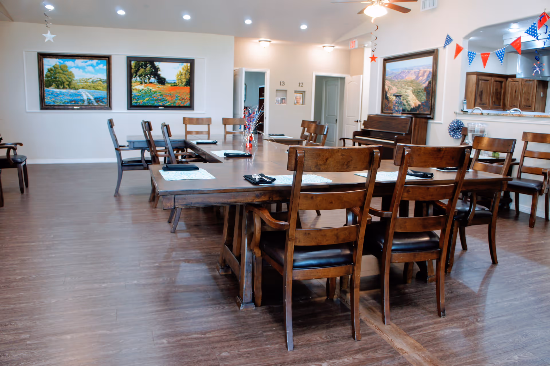 A dining room with wooden tables and chairs arranged neatly. The tables have placemats and folded napkins. The room has wooden flooring, several landscape paintings on the walls, and a piano in the corner. There are festive red, white, and blue decorations hanging near a pass-through window to the kitchen area.