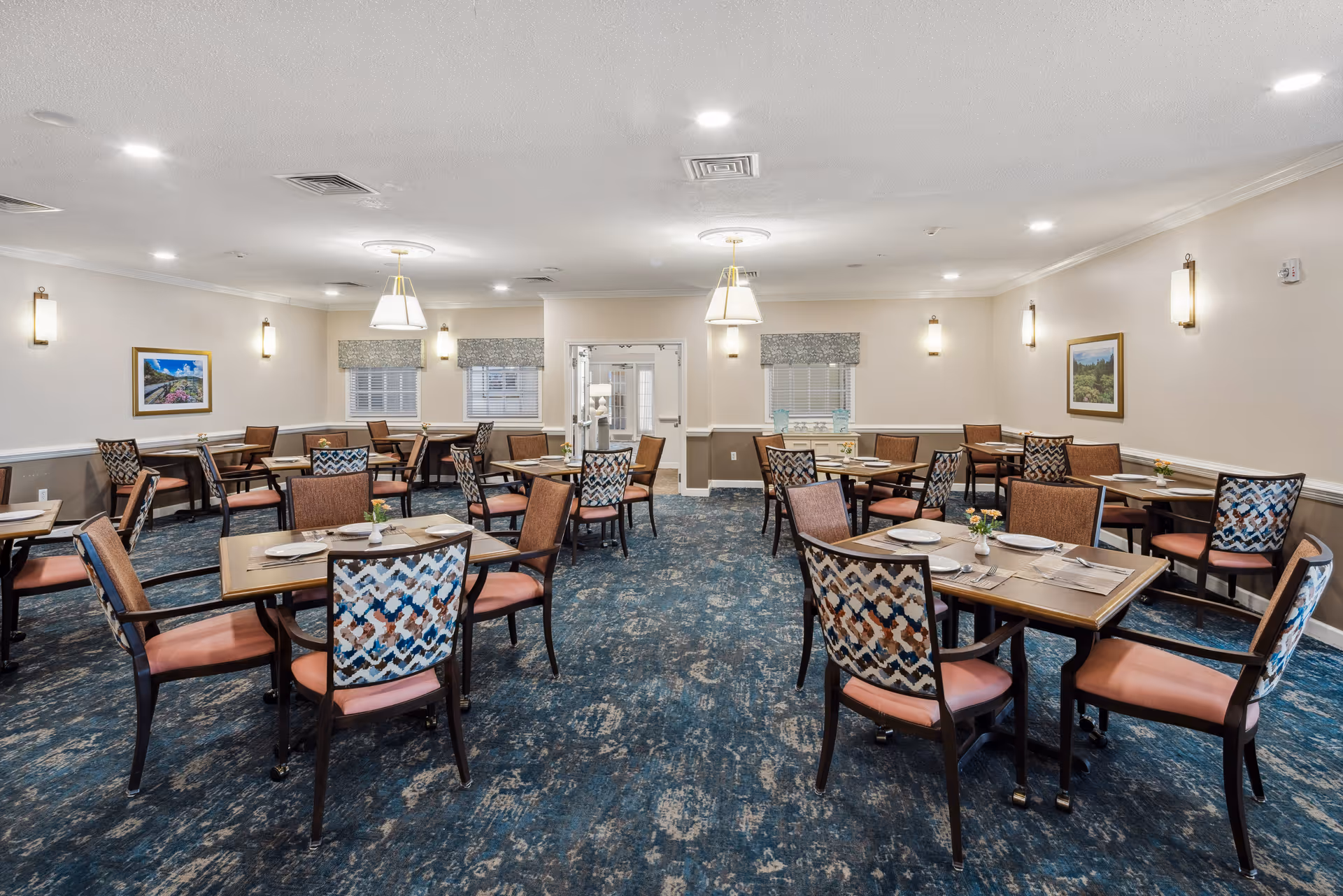 A well-lit dining room with multiple wooden tables and chairs arranged neatly. Each table is set with plates, napkins, and small flower vases. The room has blue patterned carpet, beige walls with framed landscape paintings, and several wall-mounted lights. Two pendant lights hang from the ceiling, and windows with patterned valances are visible at the far end.