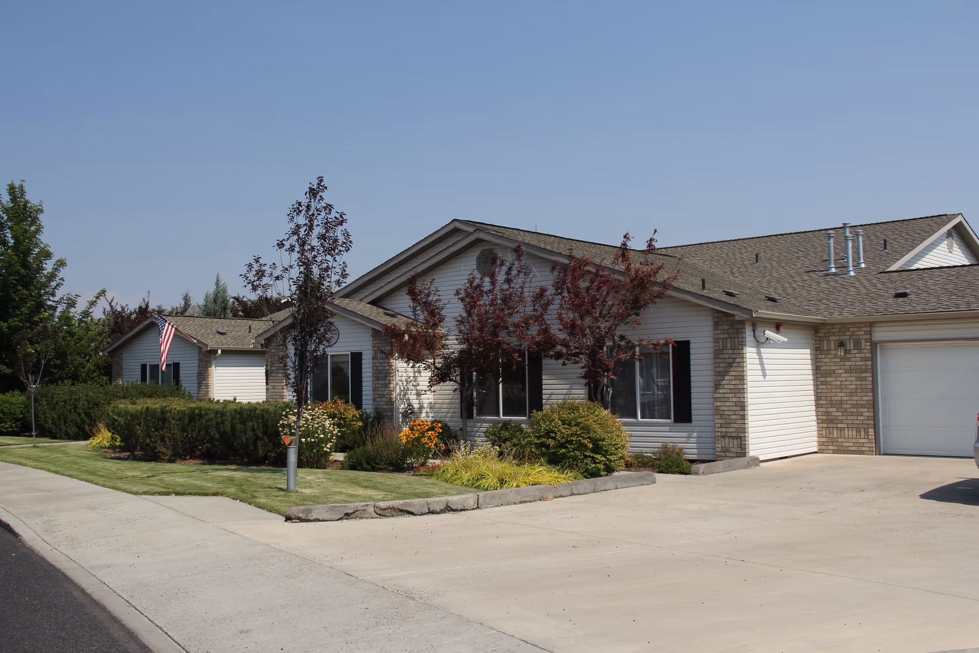 Single-story residential building with attached garage, landscaped front yard and an American flag under a clear sky.