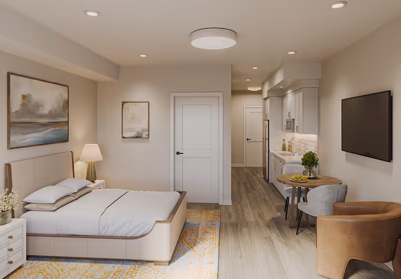 A modern studio apartment featuring a bed with white bedding and pillows, a bedside table with a lamp and flowers, two framed paintings on the wall, a small round dining table with two chairs, a brown armchair, a wall-mounted flat-screen TV, and a kitchenette with white cabinets, a sink, microwave, and refrigerator. The room has wood flooring and neutral-colored walls.