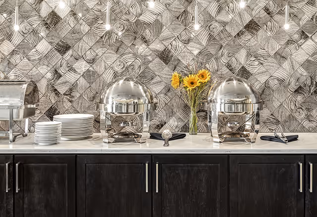 A buffet setup with two silver chafing dishes, a stack of white plates, black napkins with serving utensils, and a vase with yellow sunflowers on a white countertop. The background features a textured wall with a geometric pattern.