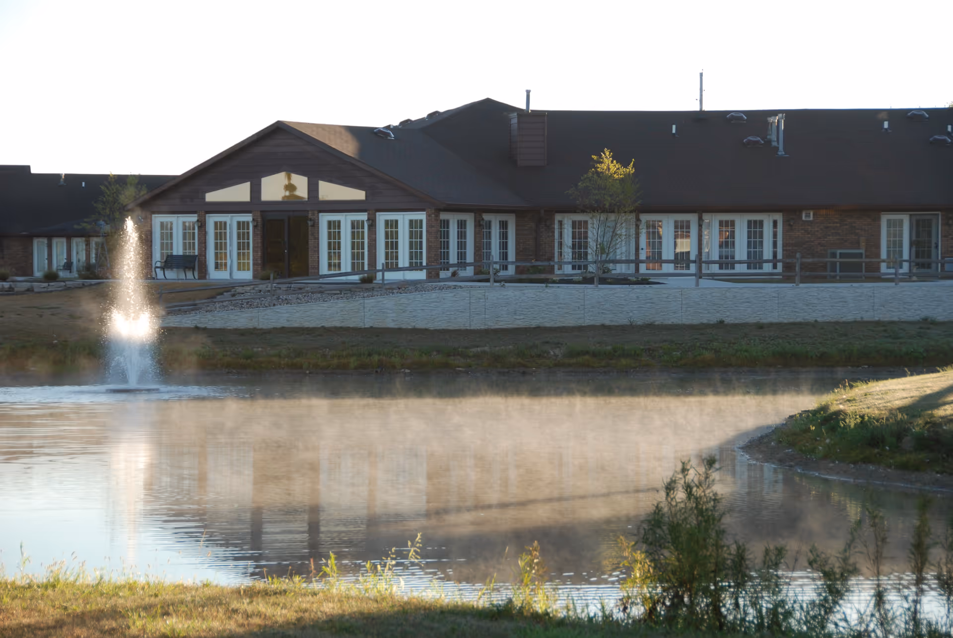 Low-rise brick health care building with multiple French doors facing a pond and a fountain in the foreground.