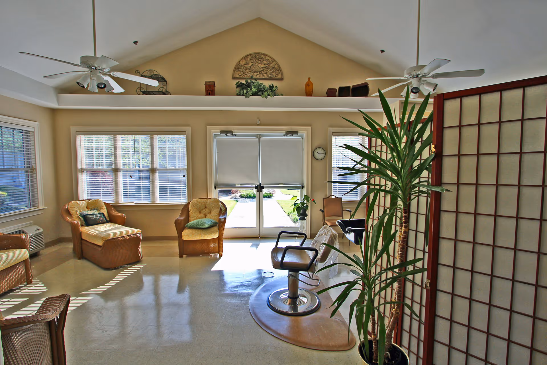 A bright room with large windows and double glass doors letting in natural light. The room features wicker chairs with cushions, a salon-style chair on a mat, ceiling fans, a tall potted plant, and a decorative folding screen. The walls are painted beige and there is a decorative shelf with plants and ornaments above the doors.