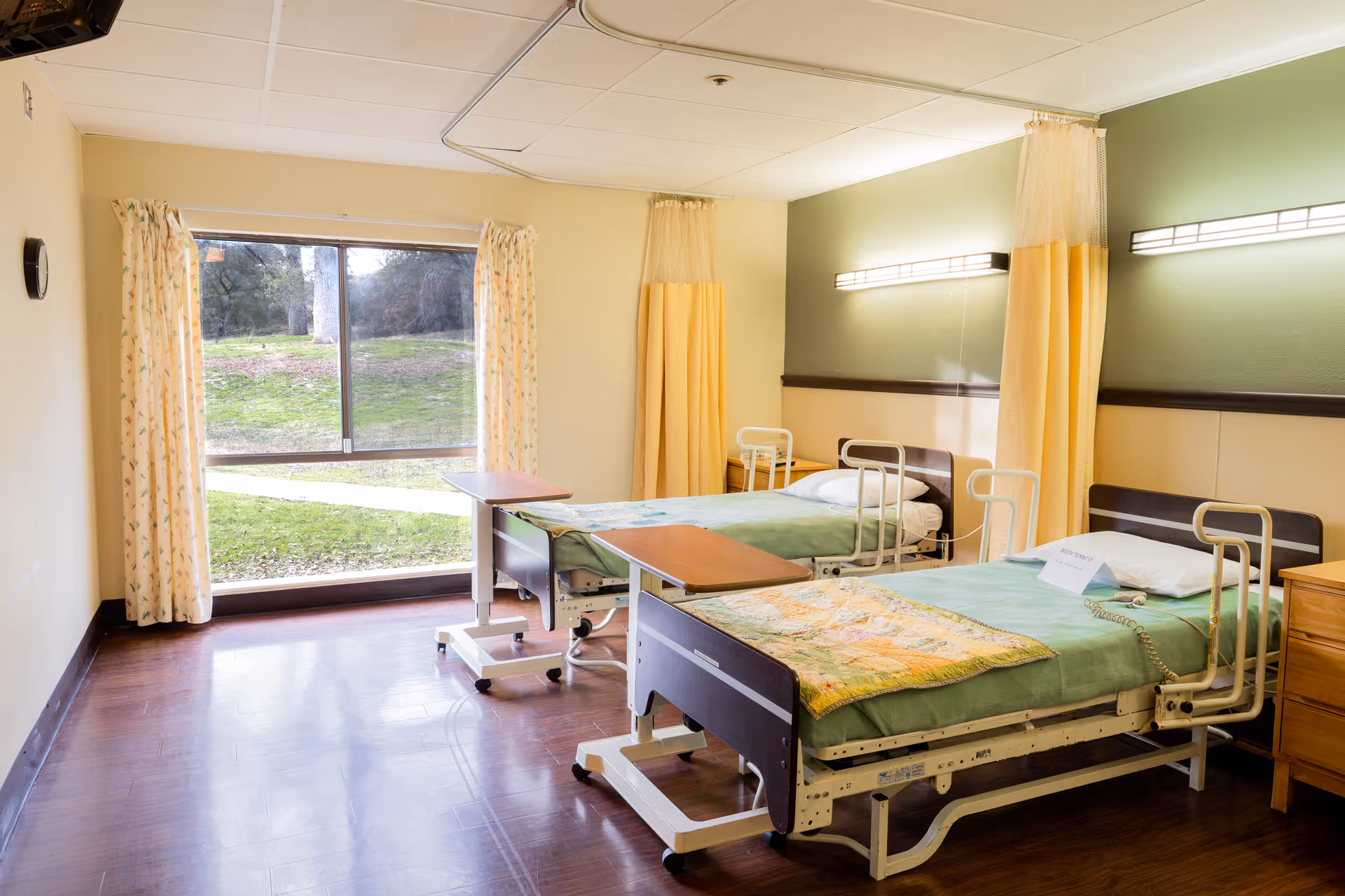 A bright room with two hospital-style beds, each with green bedding and a small overbed table. The room has large windows with curtains, letting in natural light and showing a grassy outdoor area. There are privacy curtains between the beds and wall-mounted lights above each bed.