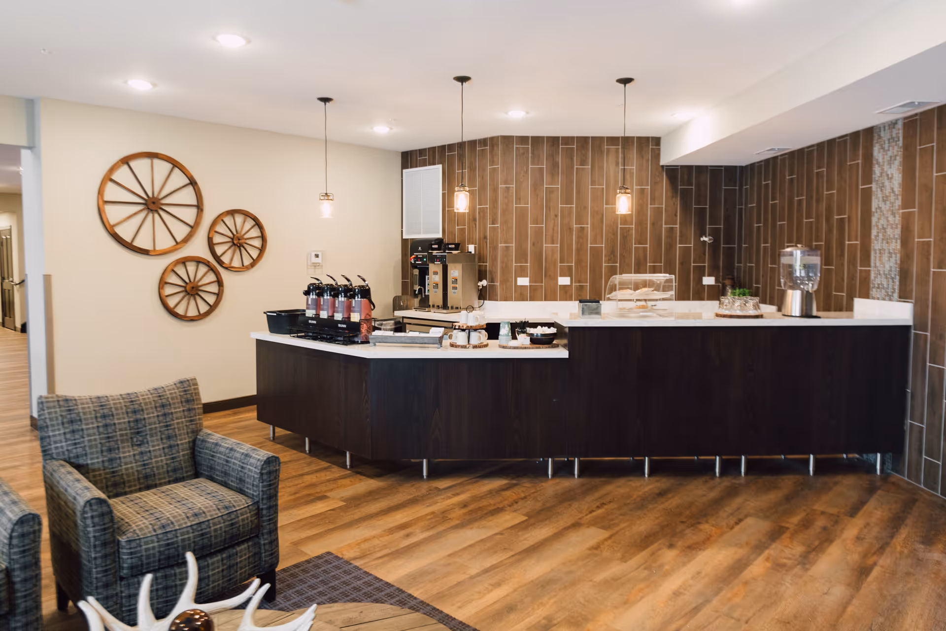 Interior view of a modern refreshment area with a dark wood counter, coffee and beverage dispensers, and a display case with pastries. The wall behind the counter is decorated with vertical brown tiles and three pendant lights hang from the ceiling. To the left, there are three decorative wooden wagon wheels mounted on a light-colored wall and a plaid upholstered armchair is partially visible in the foreground.