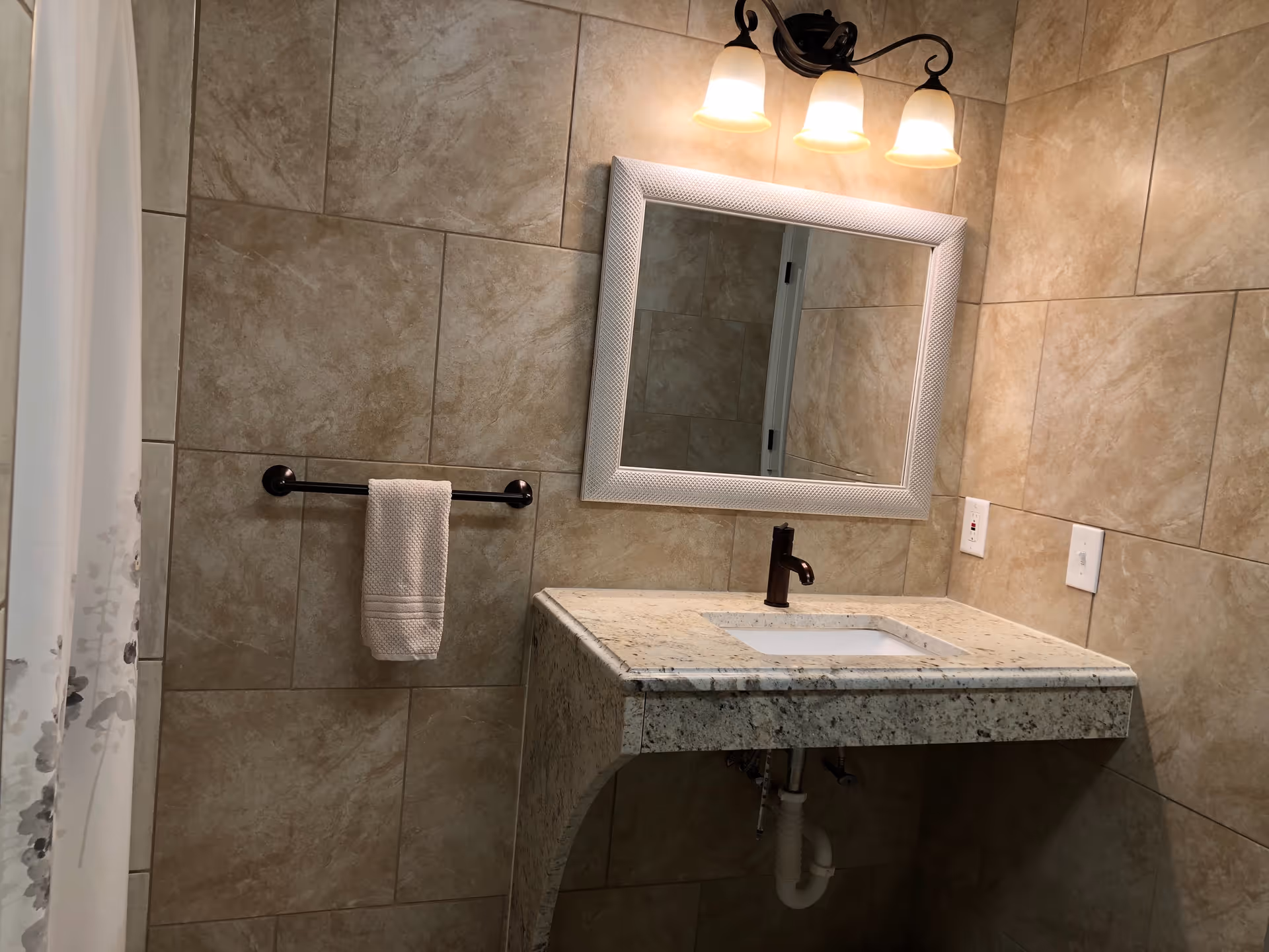 A bathroom with beige tiled walls, a granite countertop with an under-mount sink, a bronze faucet, a white framed mirror above the sink, a three-light fixture with frosted glass shades, a towel rack with a white hand towel, and a white shower curtain with a floral pattern on the left side.