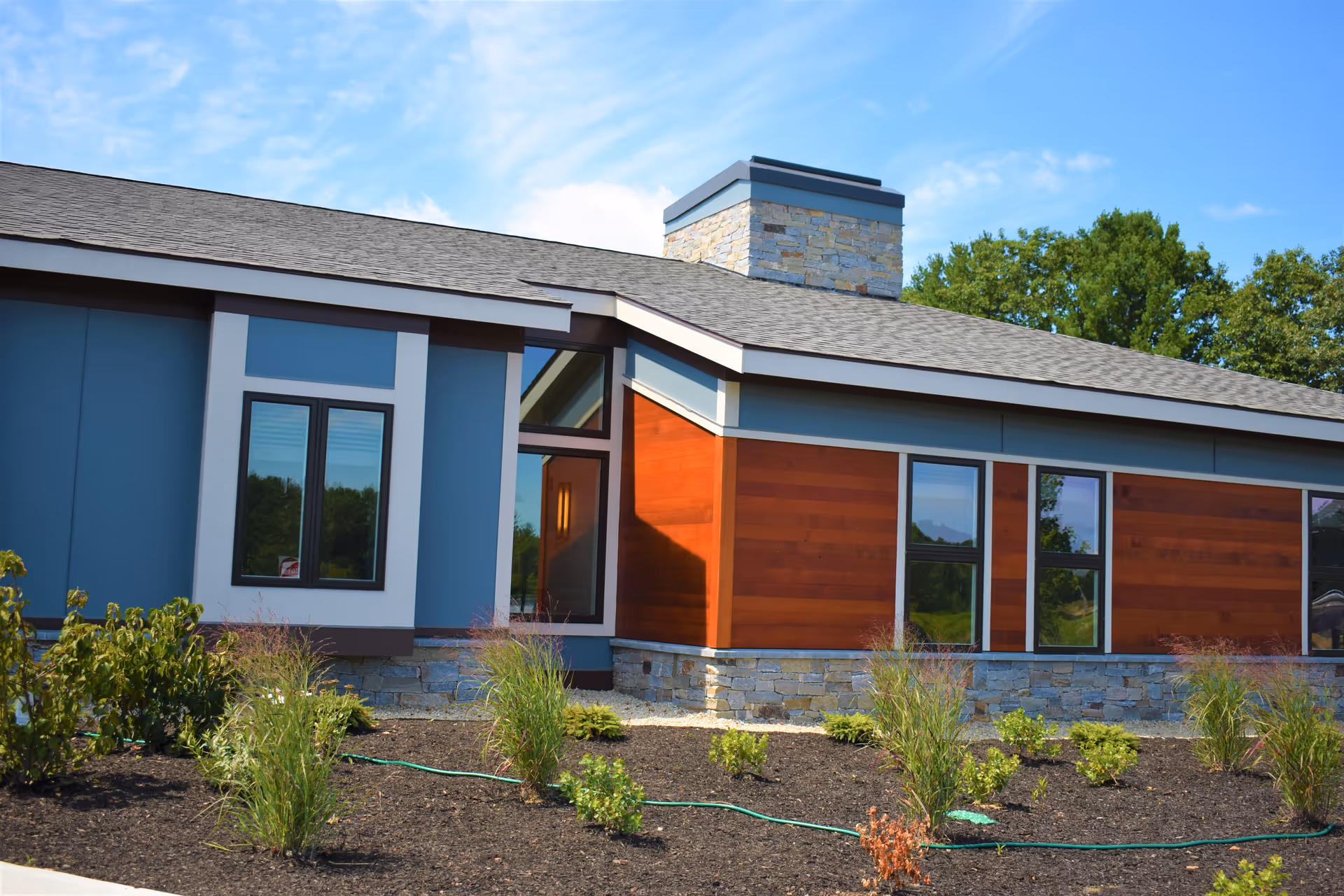 Exterior view of a modern building with a combination of blue and wooden panel siding, stone accents, and a shingled roof under a partly cloudy blue sky. There are several windows and a landscaped garden with small plants and mulch in the foreground.