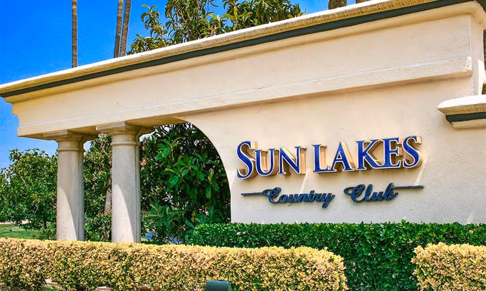 Stone entrance sign reading "Sun Lakes Country Club" flanked by columns, hedges, and palm trees.