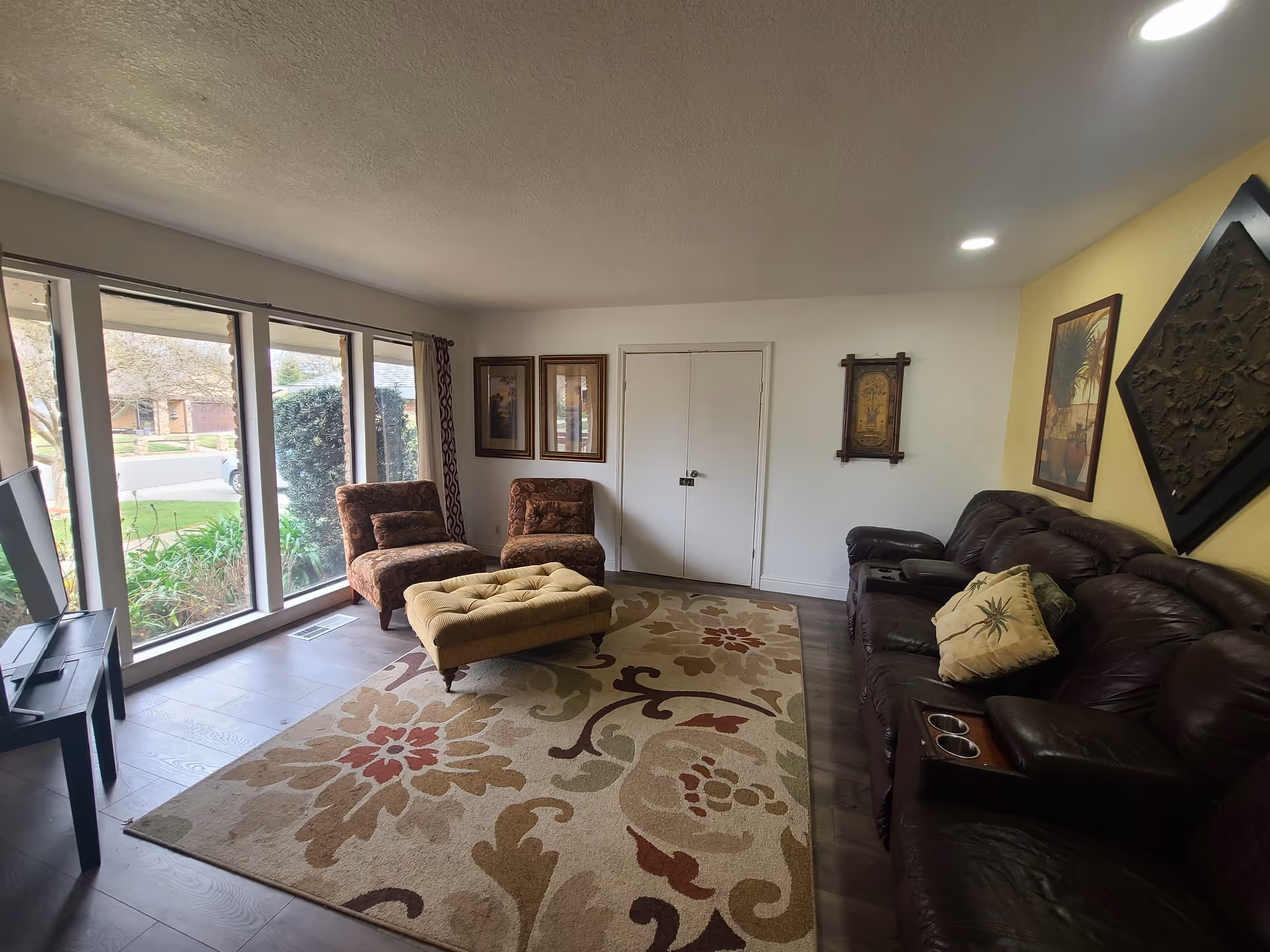A cozy living room with a large floral patterned area rug, two upholstered chairs, a tufted ottoman, and a dark brown leather sofa with a decorative pillow. The room has large windows letting in natural light, framed artwork on the walls, and a TV on a stand near the windows.