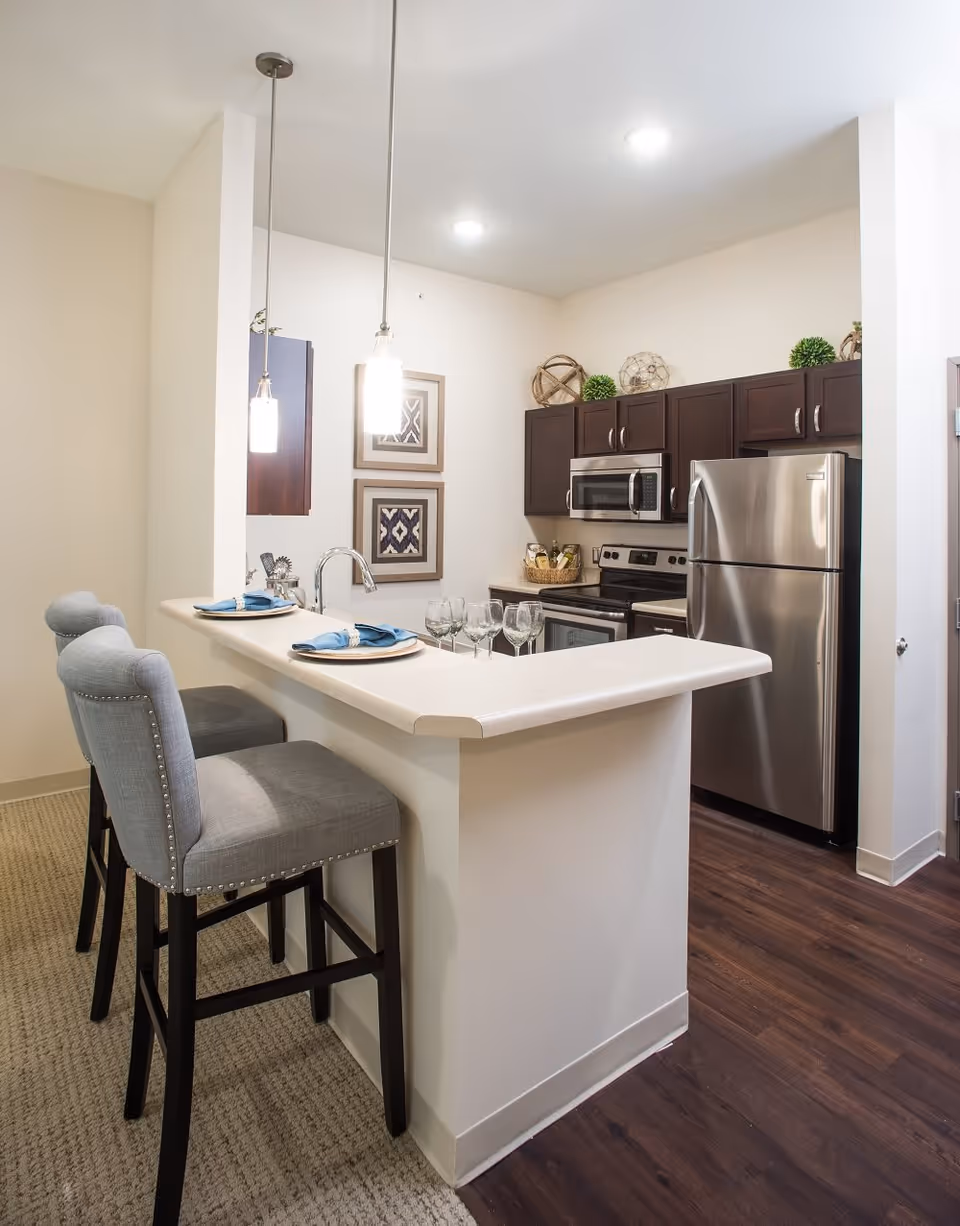 Modern kitchen area with dark wood cabinets, stainless steel refrigerator, stove, and microwave. A white countertop bar with two gray upholstered bar stools is set with plates, napkins, and wine glasses. Two pendant lights hang above the bar, and decorative framed artwork is on the wall.